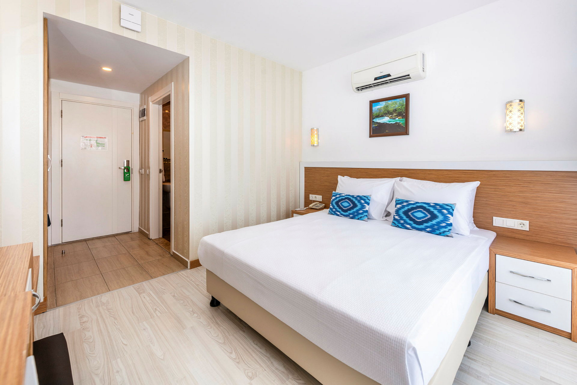Ett hotellrum med en dubbelsäng, vita sängkläder, två blåmönstrade kuddar och ett nattduksbord intill. I bakgrunden syns en dörr och ingången till ett badrum. (AI-genererad text)