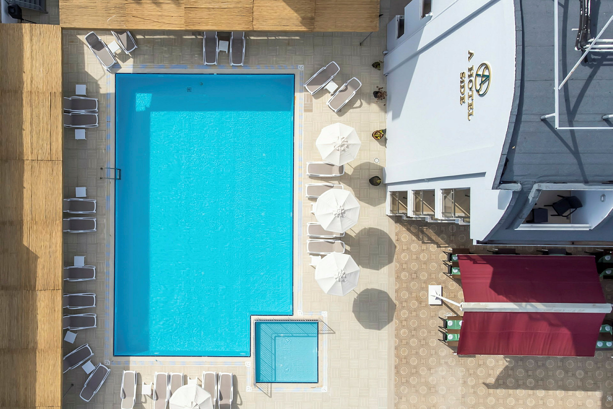 Top vy av hotellpool med solstolar och parasoller, omgiven av byggnader. (AI-genererad text)