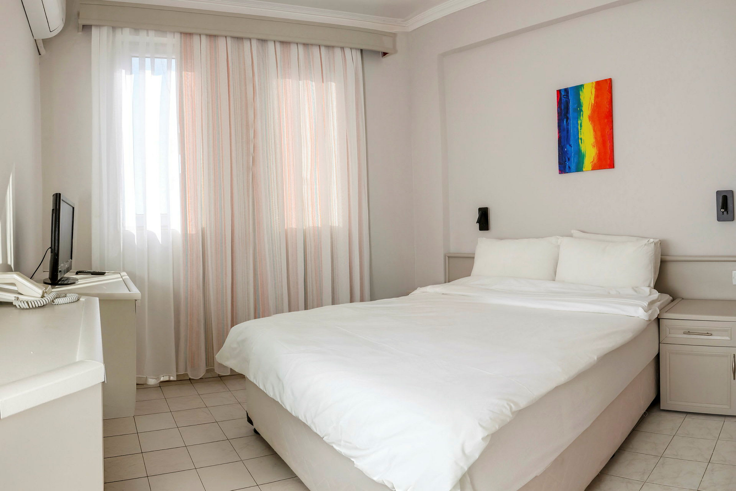 Ett hotellrum med en dubbelsäng. Väggarna är ljusa, och rummet har ett färgglatt konstverk ovanför sängens huvudände. Det finns också en liten tv och ett skrivbord vid sidan. (AI-genererad text)