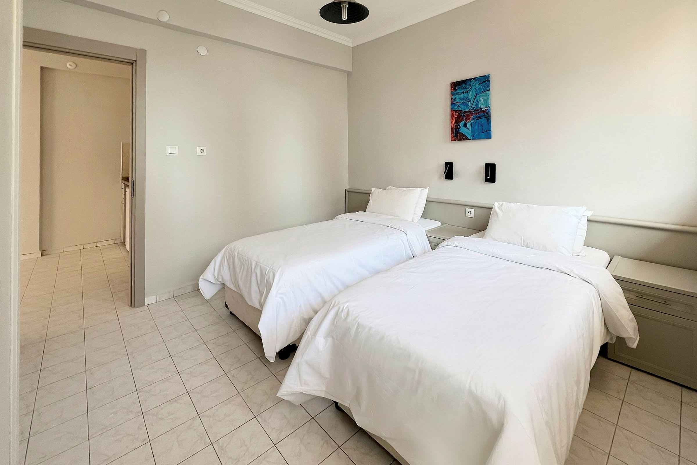 Ett hotellrum med två enkelsängar, vita sängkläder och ett ljust kaklat golv. Det finns vägglampor och en tavla på väggen ovanför sängarna. (AI-genererad text)