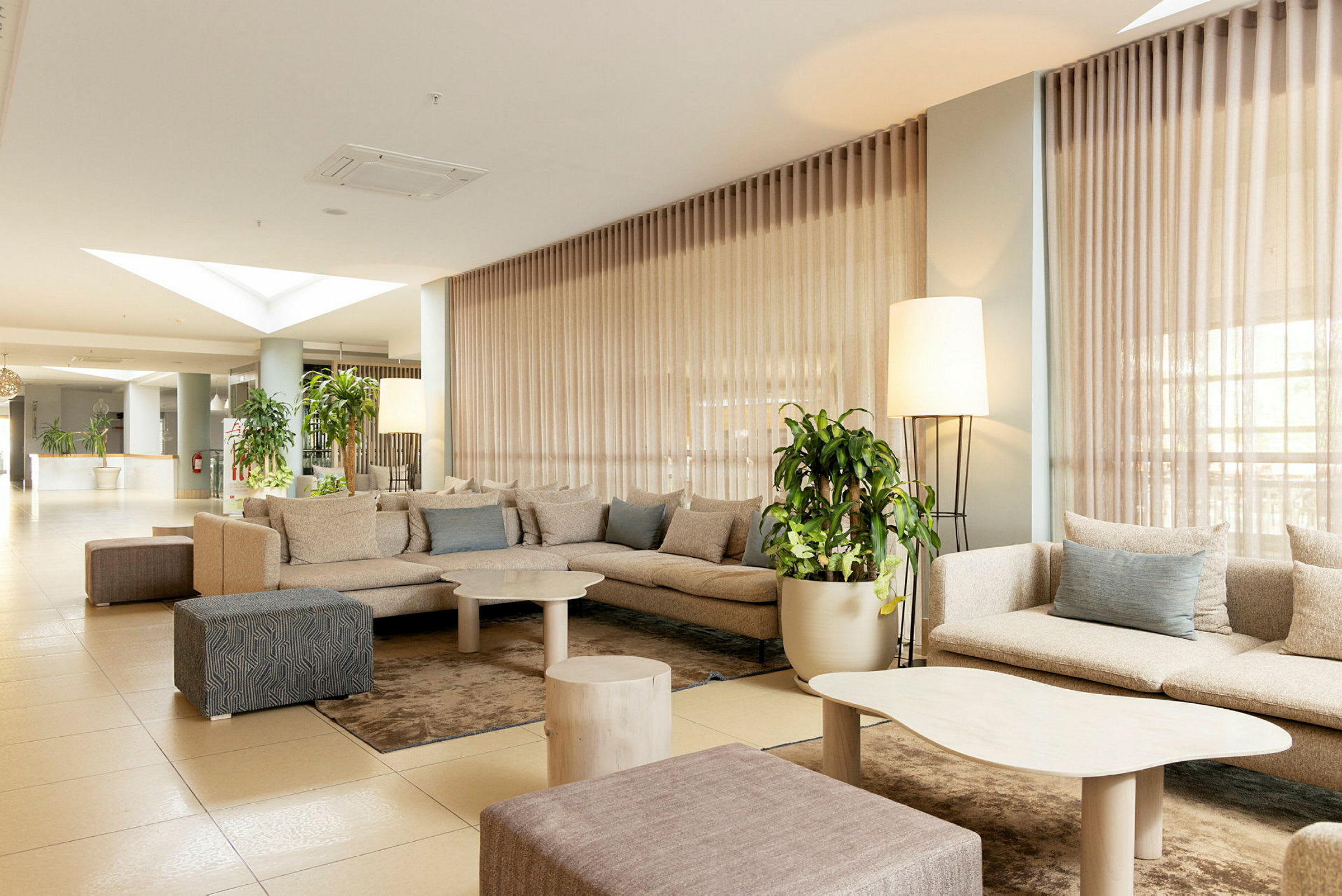 Ett hotellrum med modern design, innehåller en grupp bekväma beige soffor, låga bord och gröna växter. (AI-genererad text)