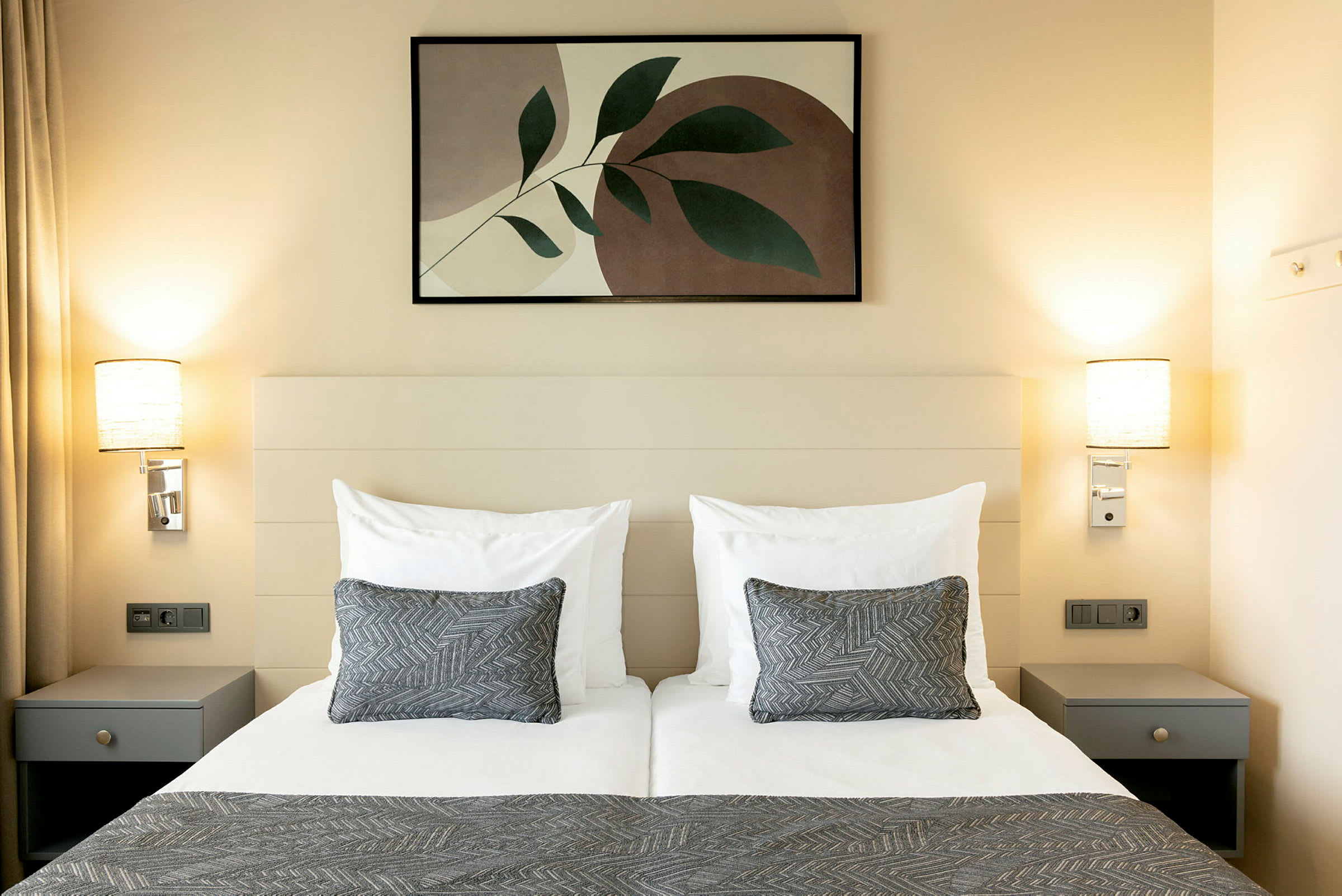 Ett hotellrum med en dubbelsäng, vita sängkläder, två grå prydnadskuddar och abstrakt väggkonst ovanför sängen. (AI-genererad text)