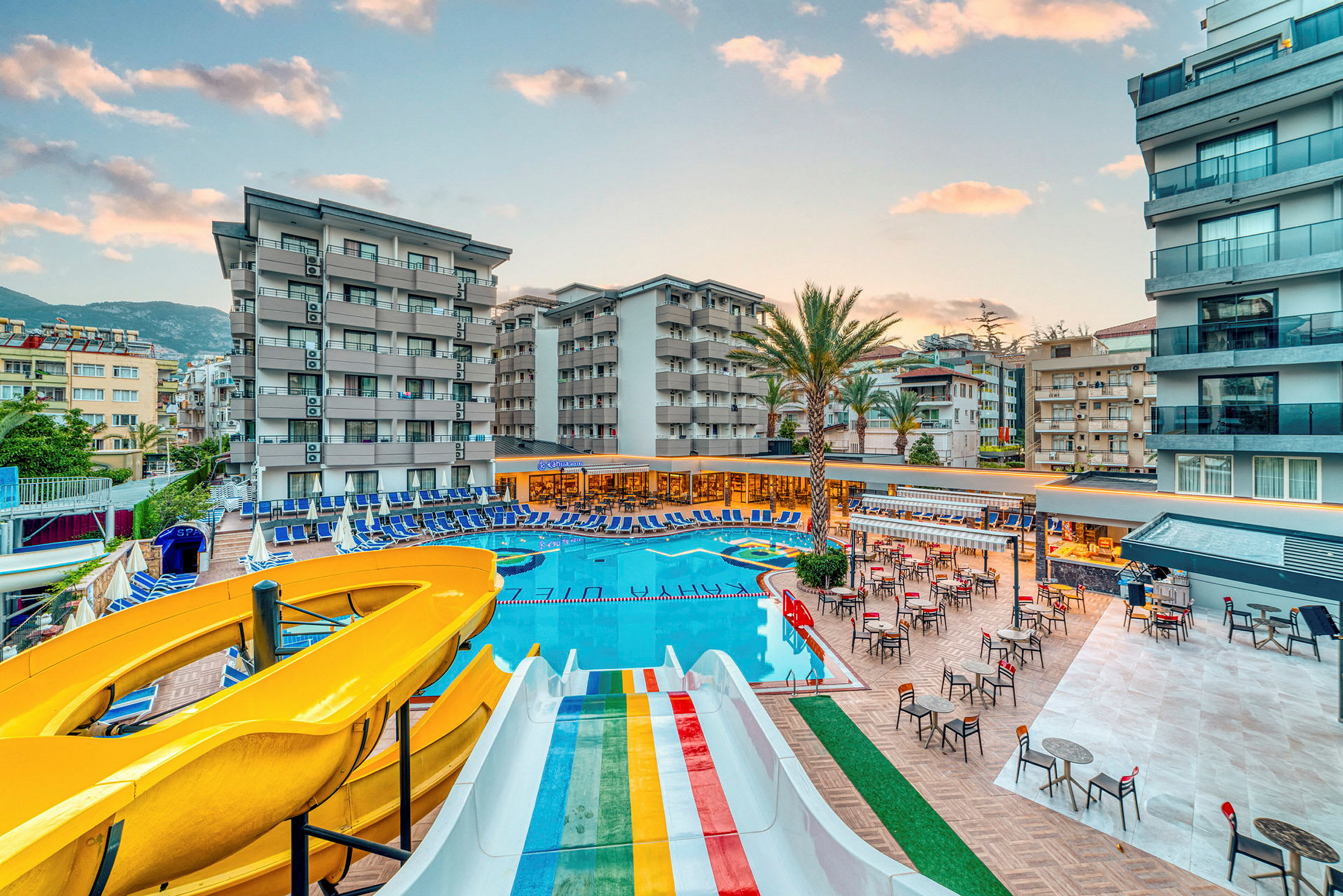 Stor gul och regnbågsfärgad vattenrutschkana vid en pool omgiven av hotellets byggnader och solstolar. (AI-genererad text)