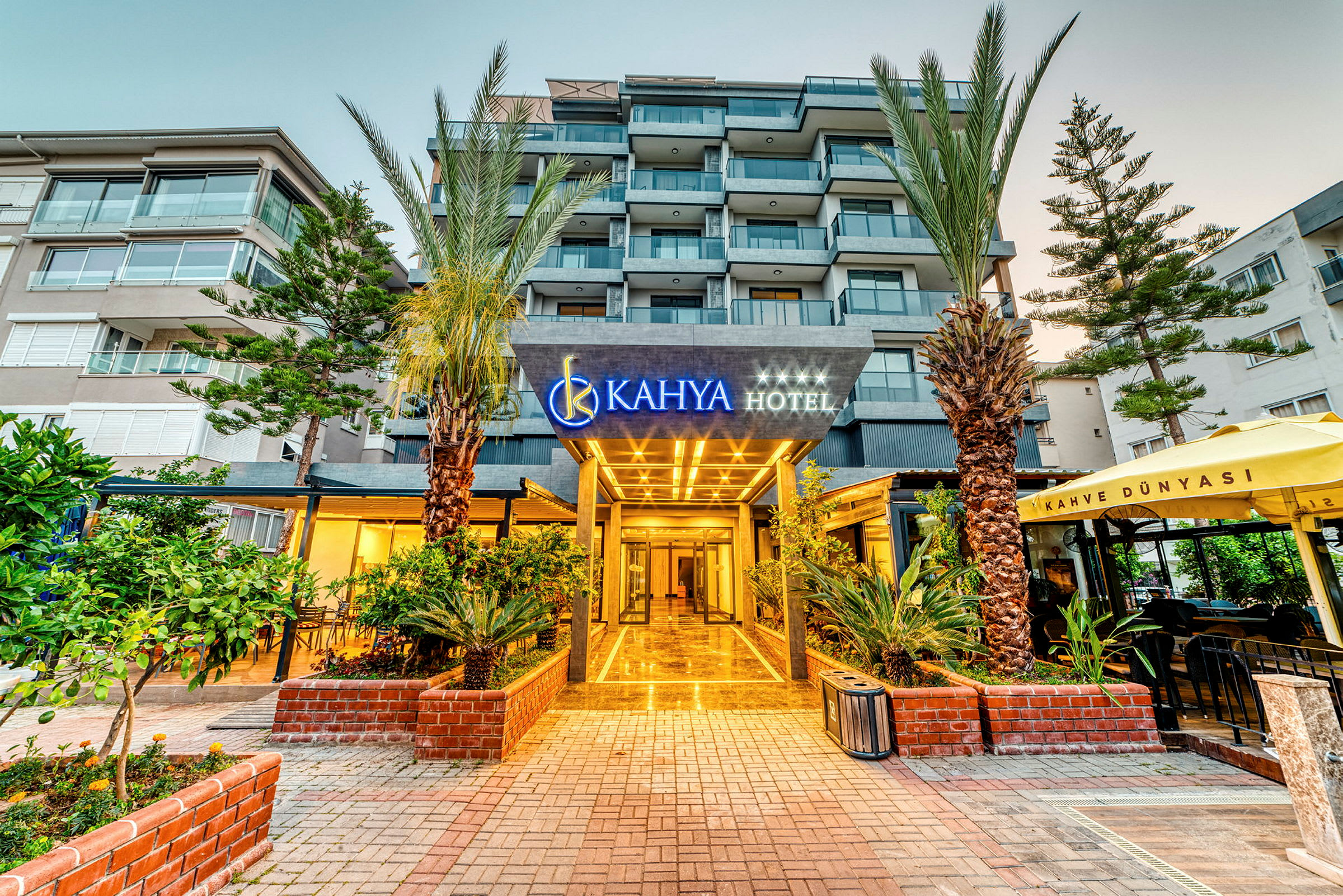 Ingång till Kahya Hotel med palmer och träd framför byggnaden. (AI-genererad text)