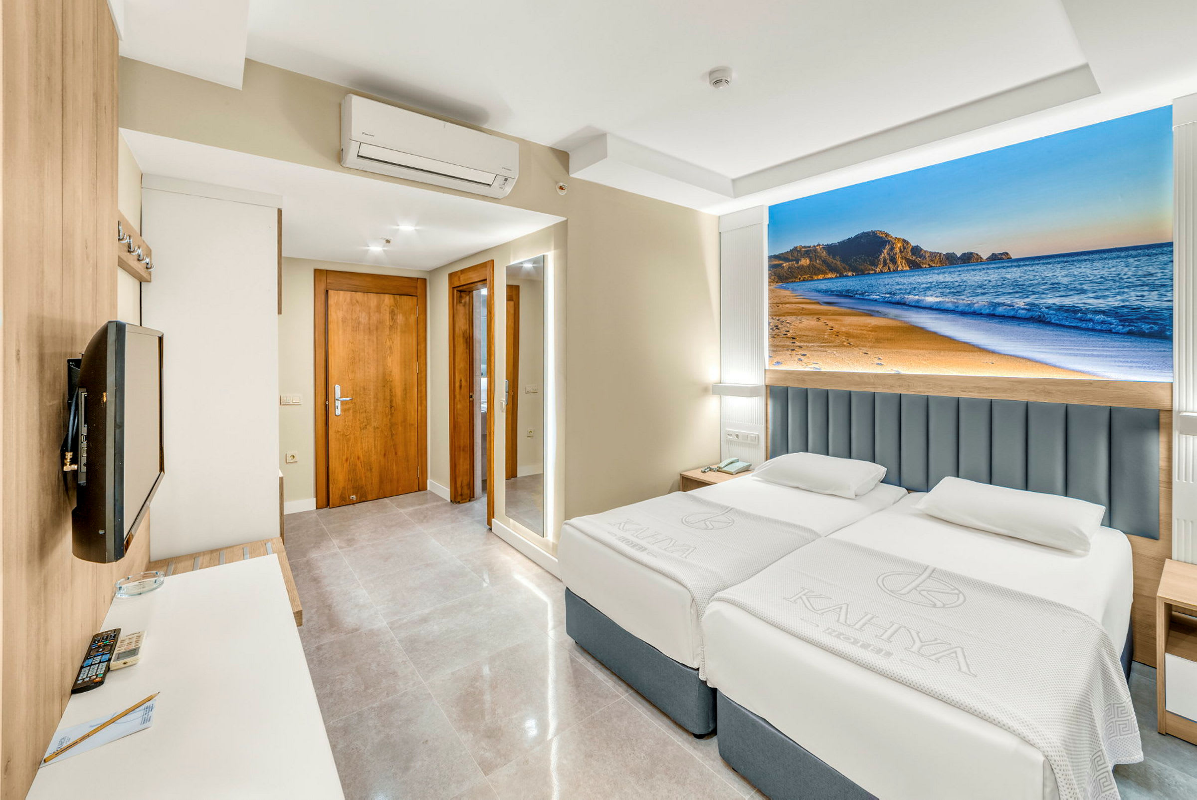 Hotellrum med två bäddar, en väggmonterad TV och en dekorativ bild av en strand bakom sängarna. (AI-genererad text)
