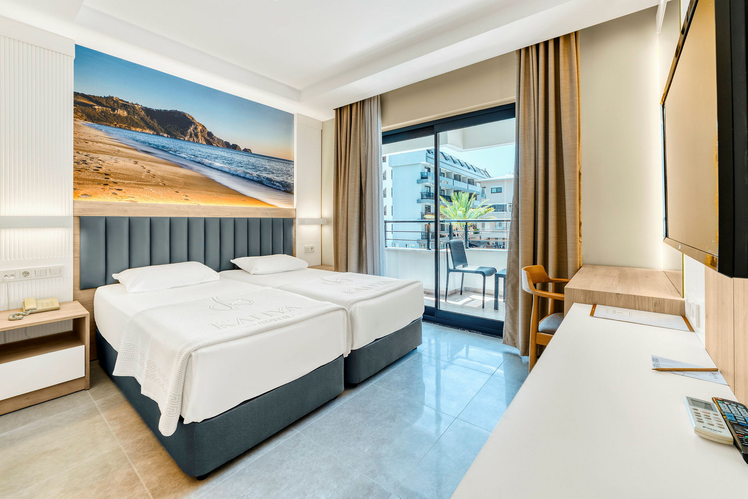 Hotellrum med två enkelsängar, balkong i bakgrunden och stort fotografi av en strand på väggen. (AI-genererad text)