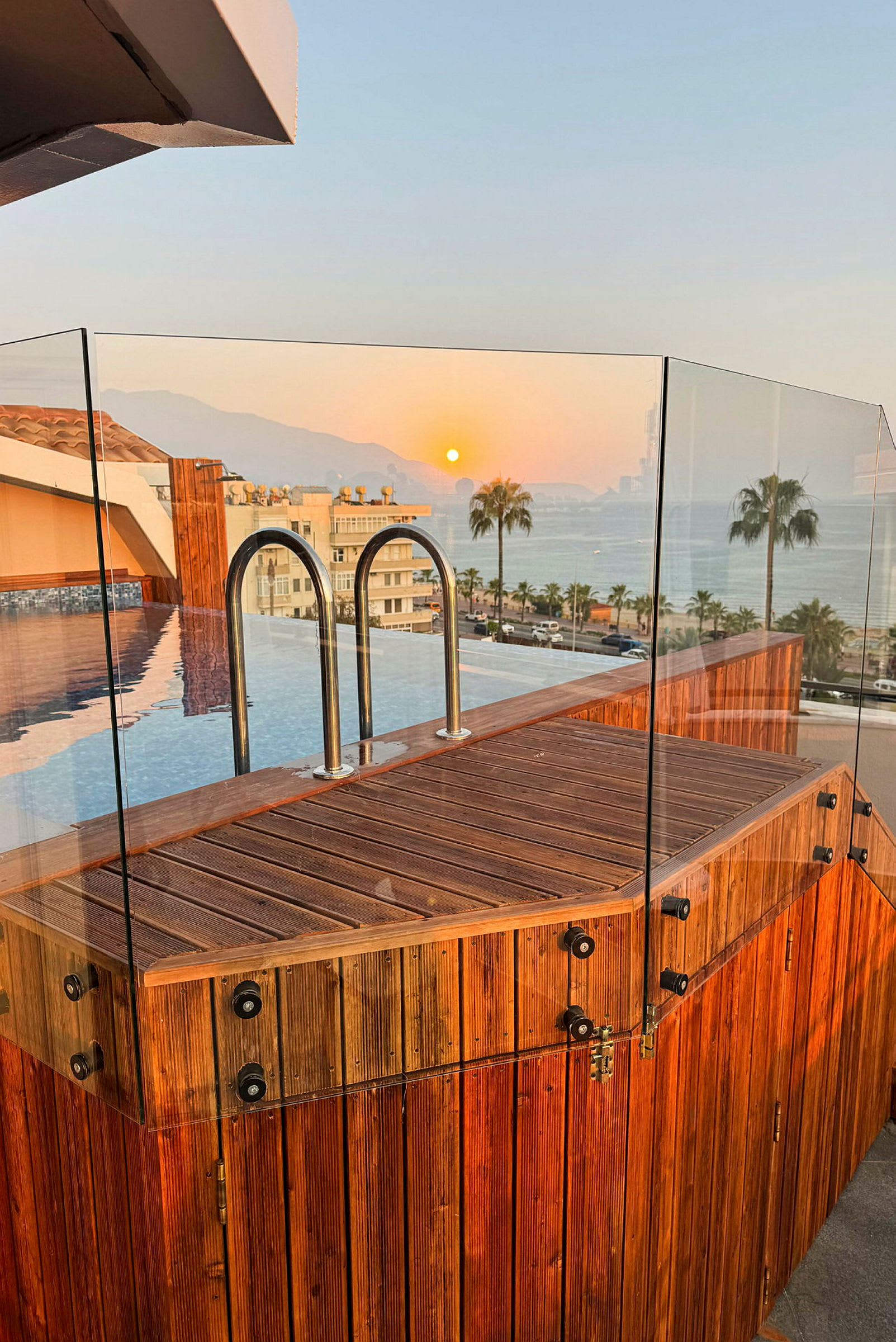 Bild av en hög belägen infinitypool omgiven av träräcken och glas, med utsikt över havet och palmer vid solnedgång. (AI-genererad text)