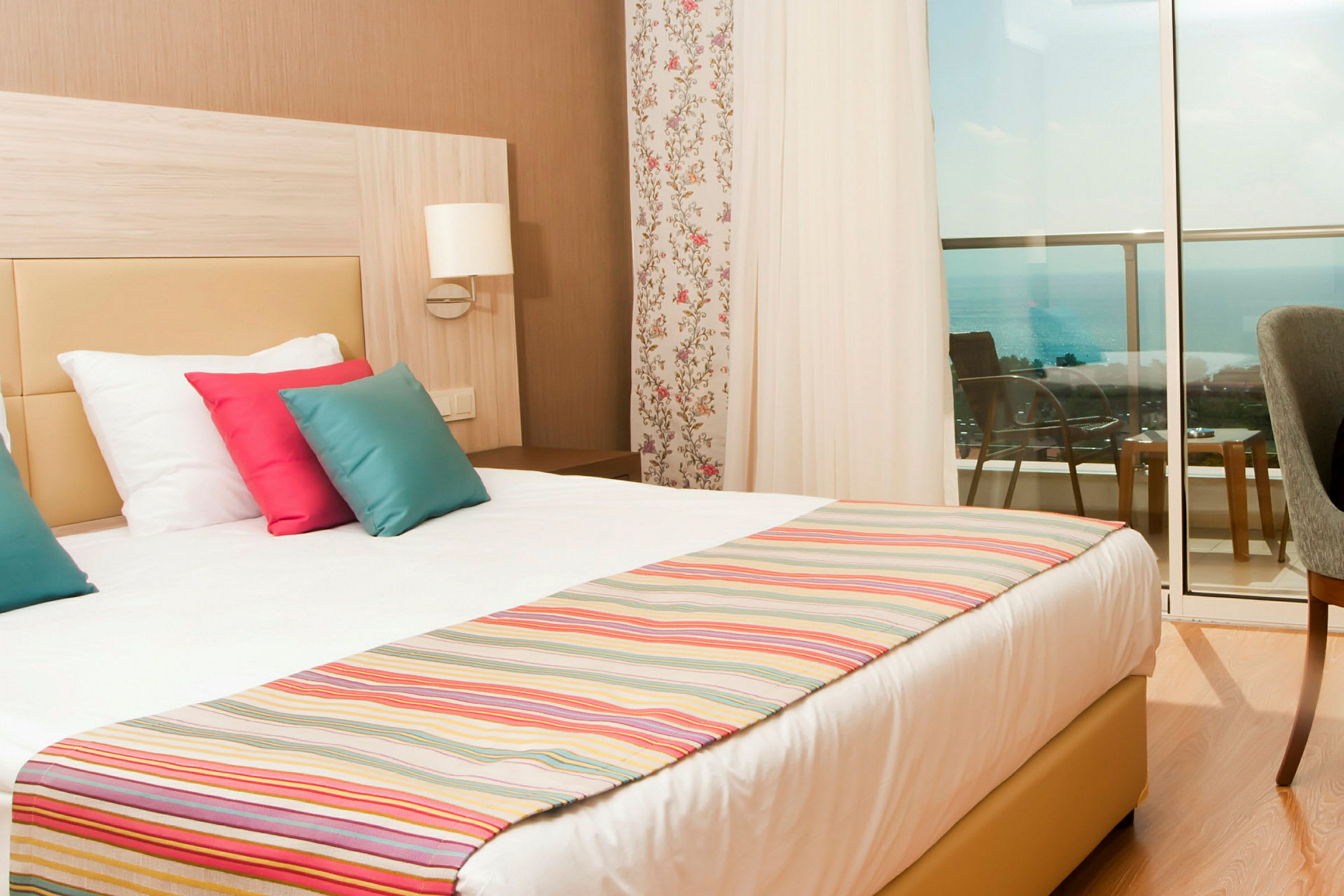 Hotellrum med en dubbelsäng, färgglada kuddar och utsikt mot havet från balkongen. (AI-genererad text)