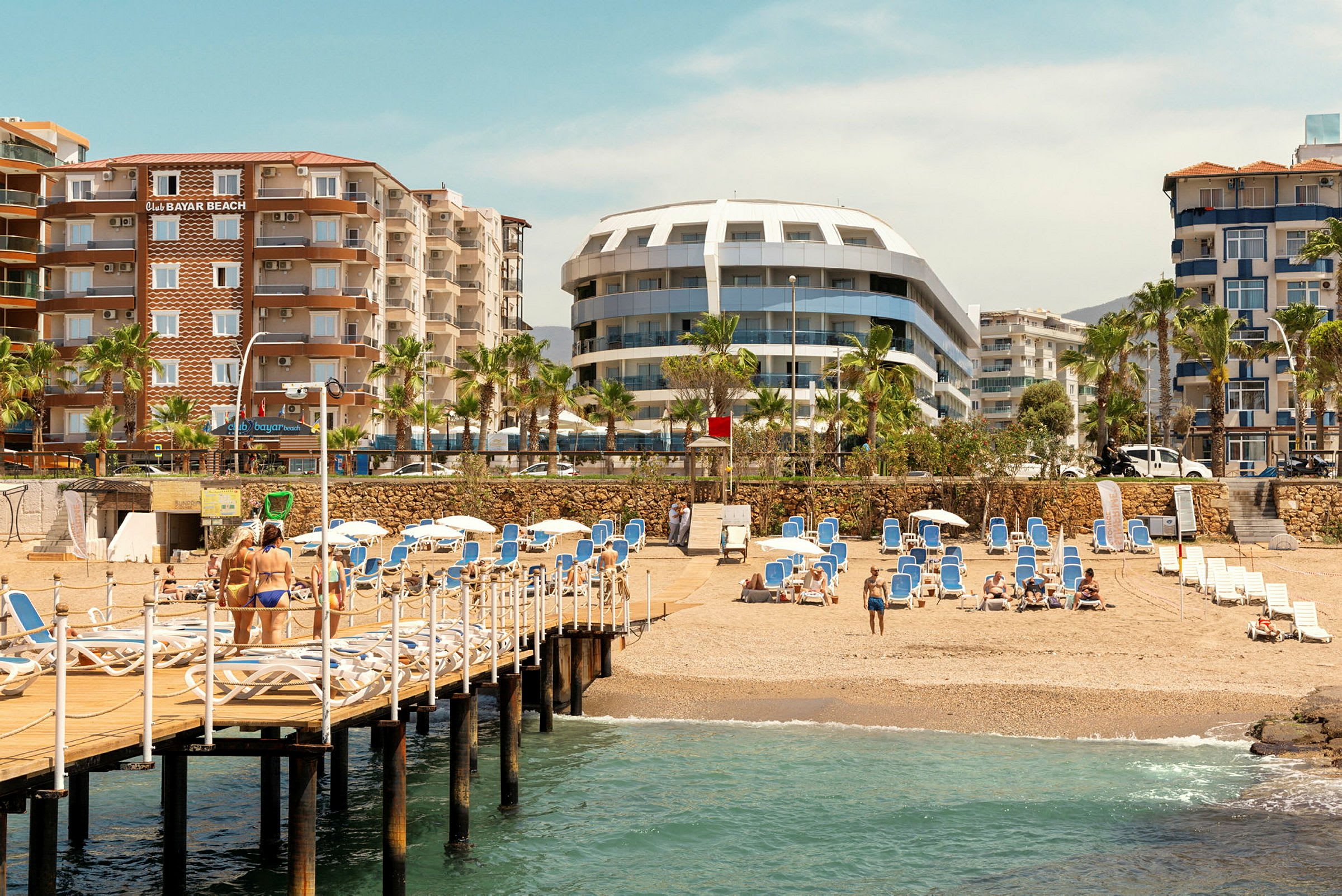 Strand med sand, solstolar, träbrygga och hotell i bakgrunden. (AI-genererad text)