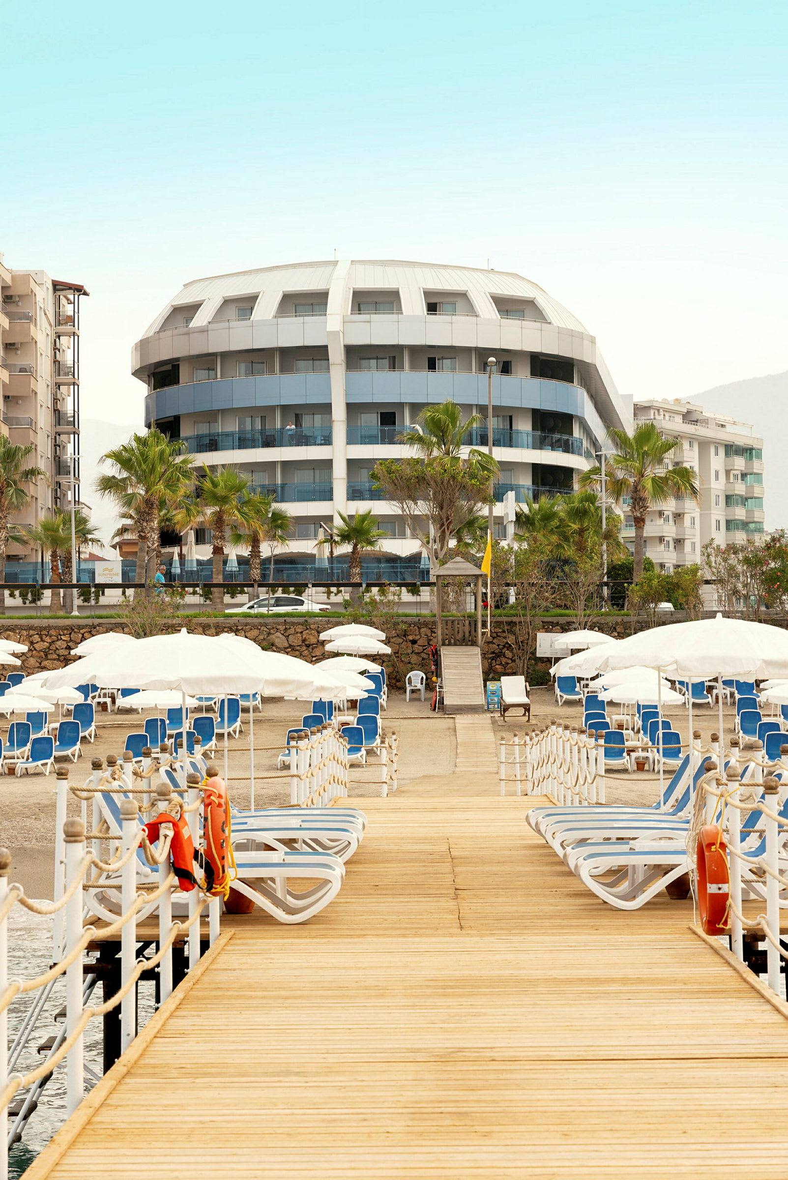 En träpir leder till solstolar och parasoller vid en strand, med ett hotell i bakgrunden. (AI-genererad text)