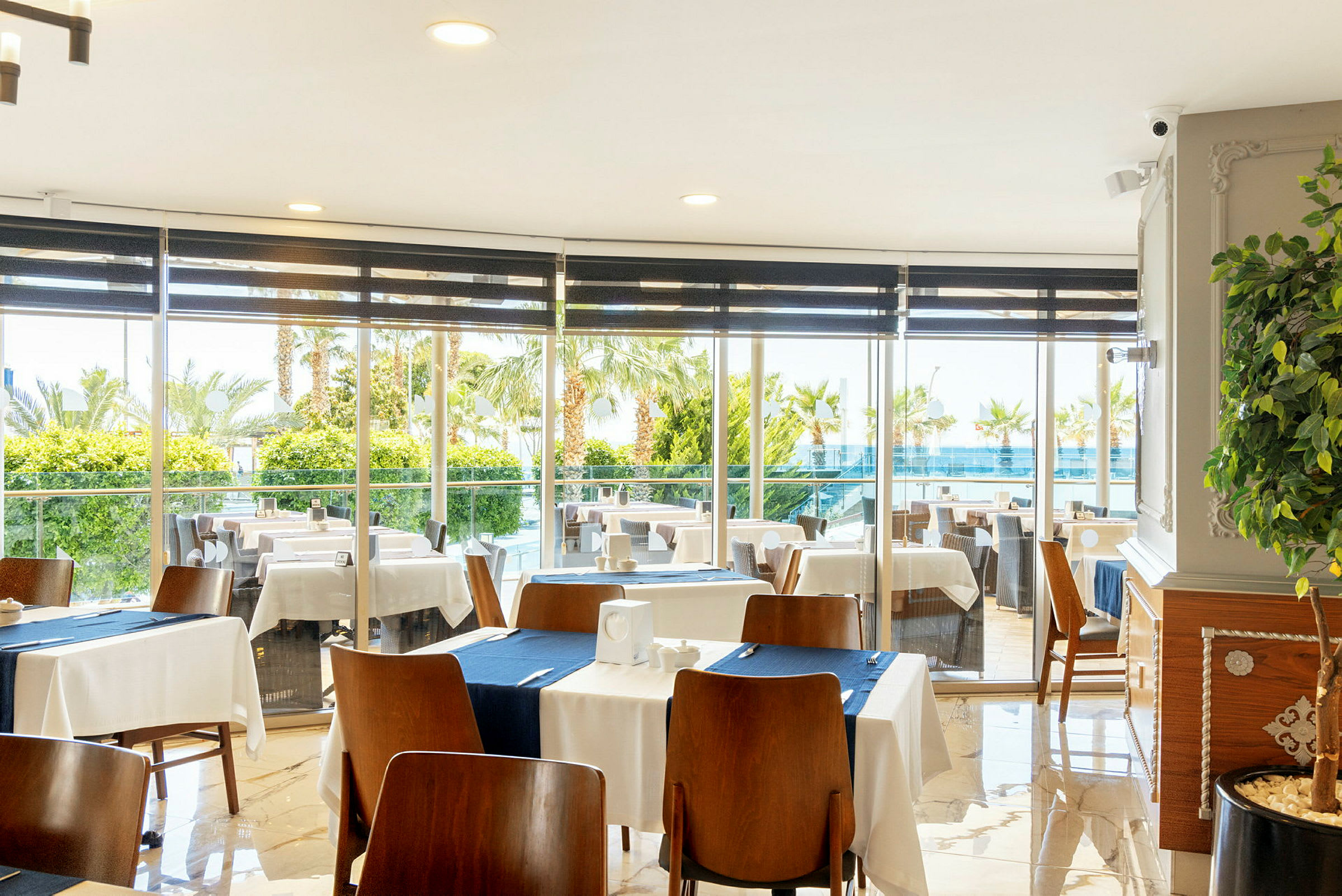 Restaurang med dukade bord och utsikt genom fönster mot grönska och pool. (AI-genererad text)