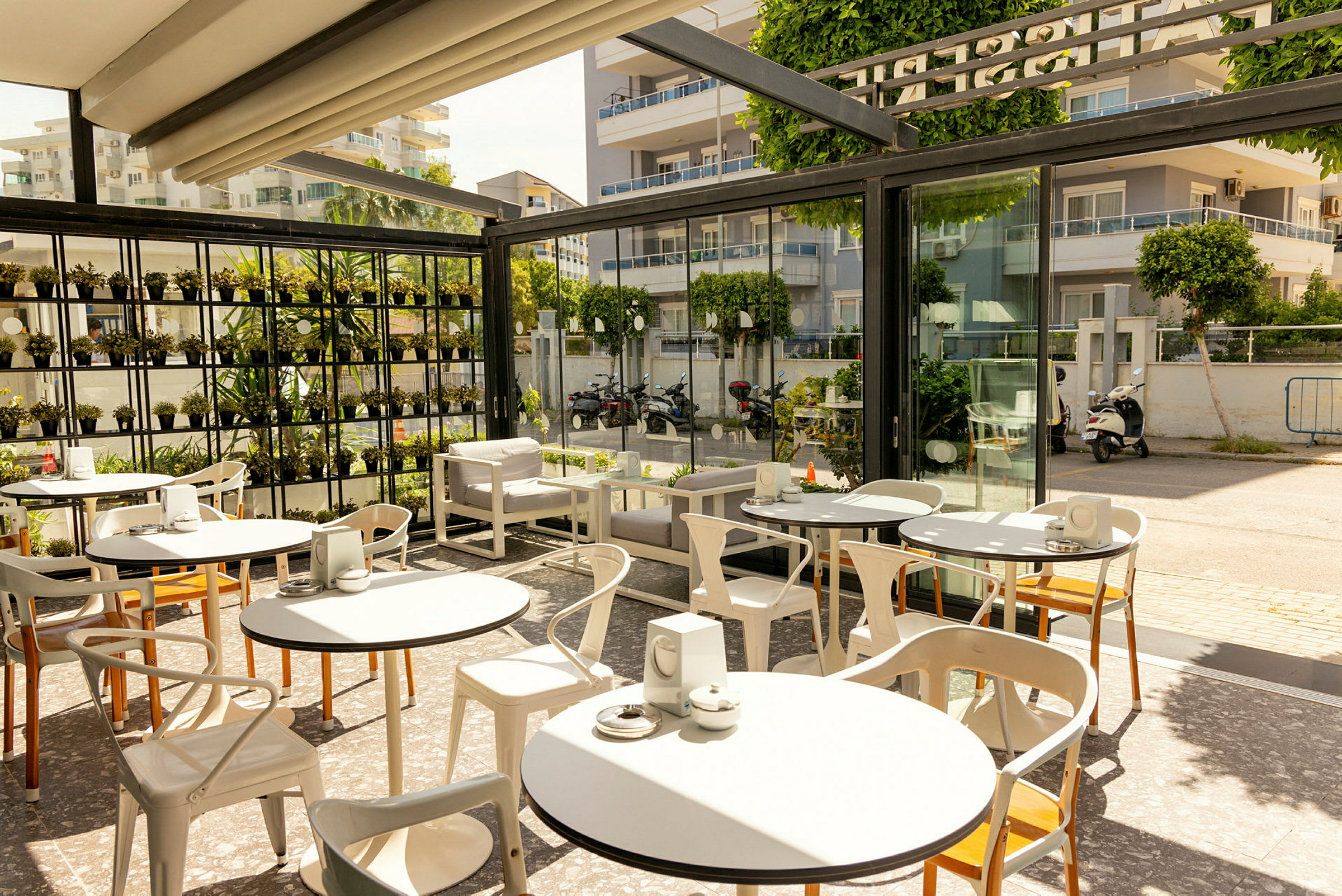 En fin caféterrass med bord och stolar, omgiven av glasväggar och gröna växter. (AI-genererad text)