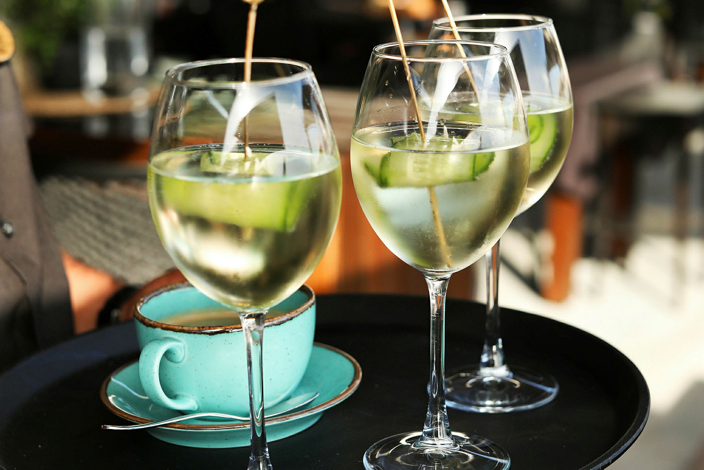 Tre glas med drinkar innehållande is och gurkskivor samt en turkos kaffekopp på ett serveringsfat. (AI-genererad text)