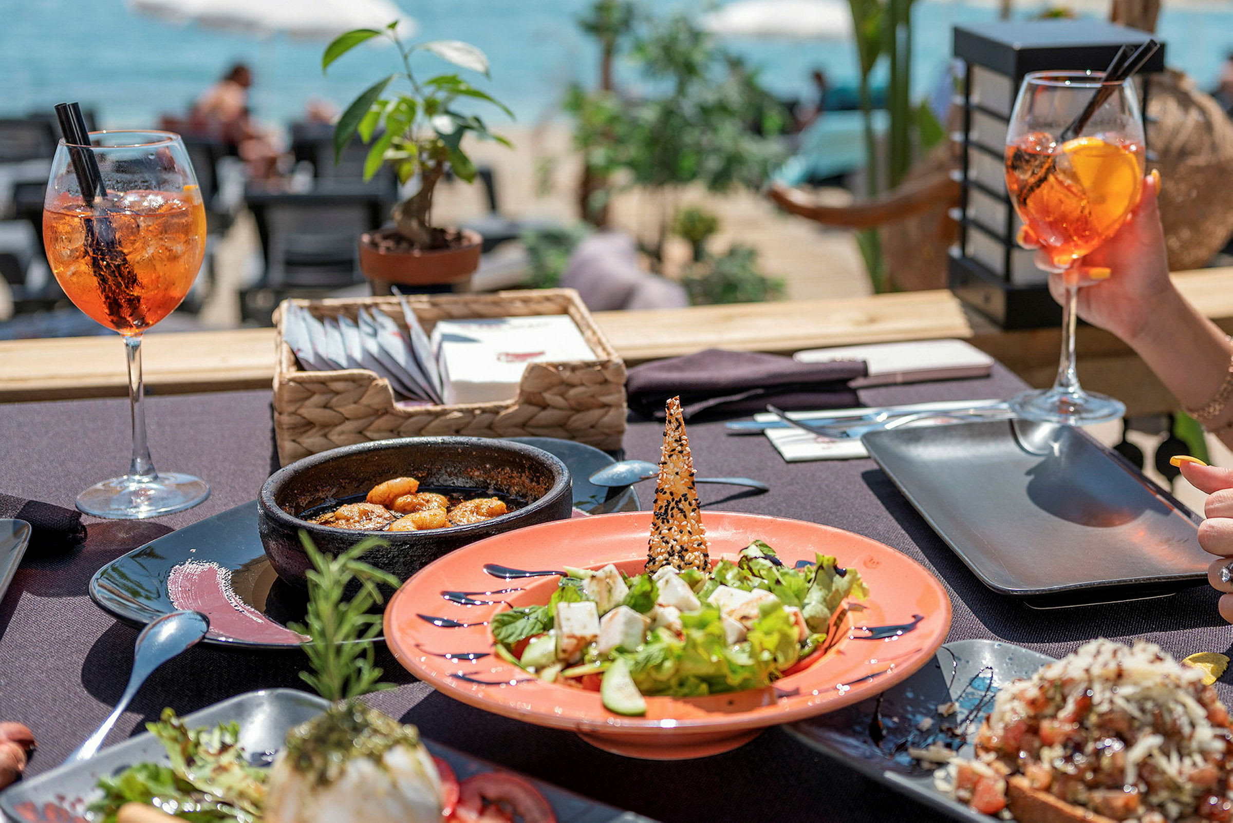 En dukad bordsuppsättning med en mängd olika maträtter och drinkar på en utomhusrestaurang vid havet. (AI-genererad text)