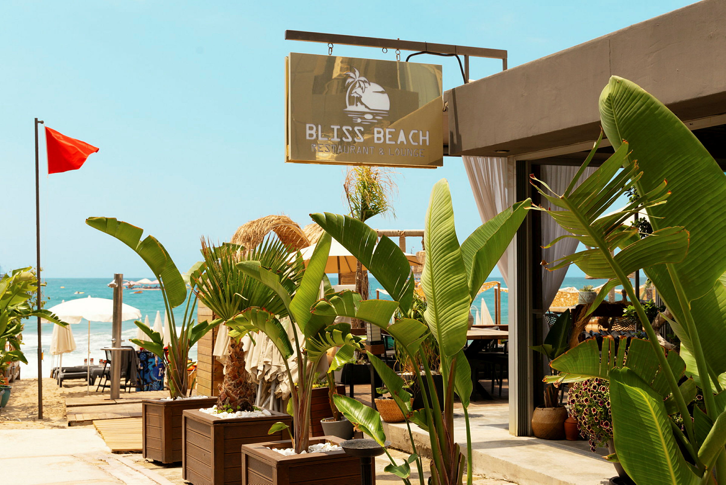 Skylt med texten Bliss Beach Restaurant & Lounge vid en strand med röda flaggor och parasoller. (AI-genererad text)