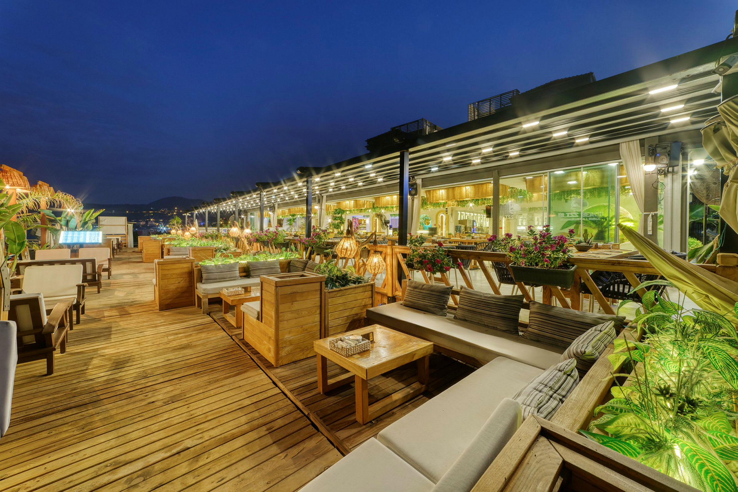 En utomhusrestaurang med träterrass, soffor och bord, samt gröna växter och ljusslingor. (AI-genererad text)