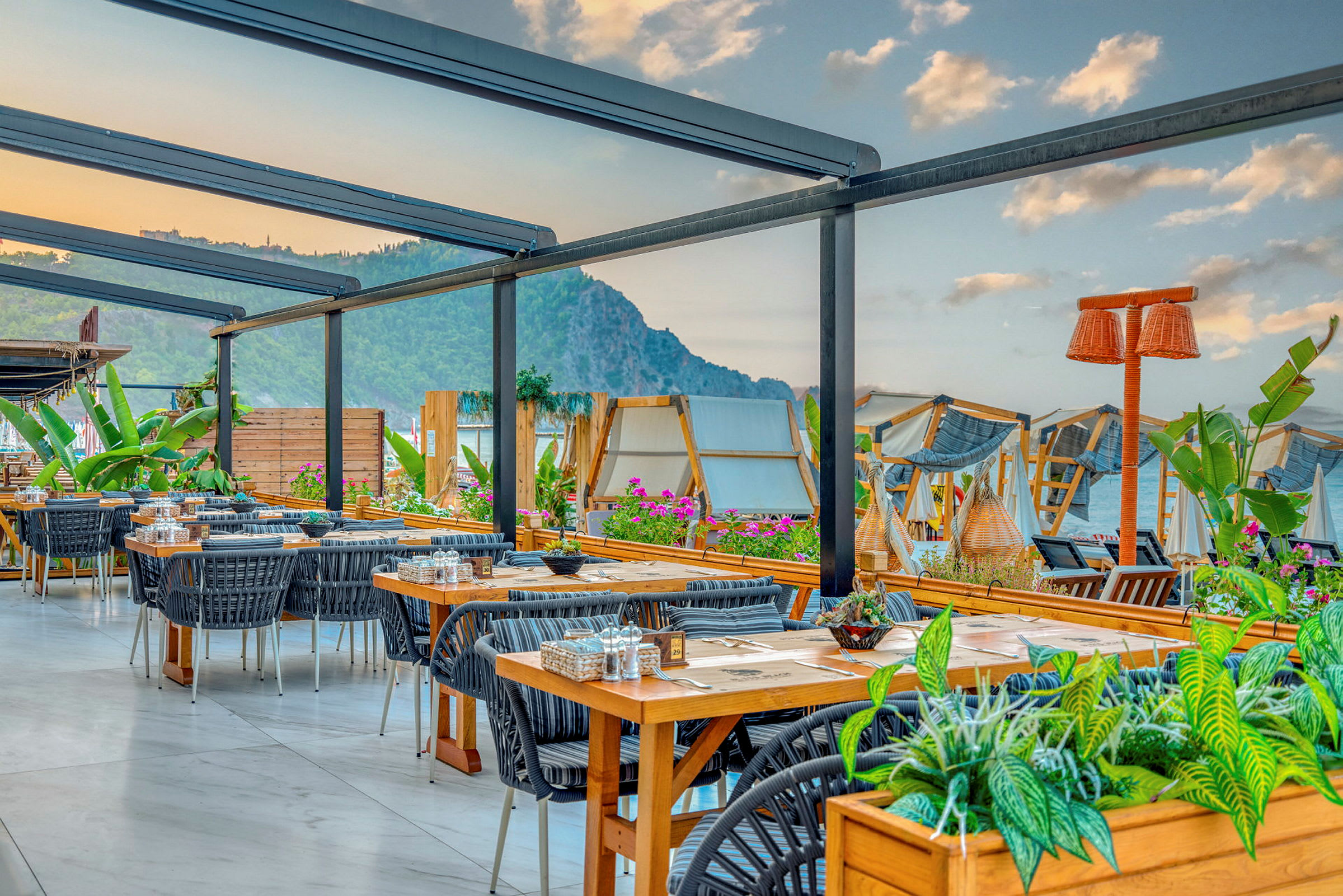 Utomhusrestaurang med bord och stolar, omgiven av växter och en utsikt över havet och bergen. (AI-genererad text)