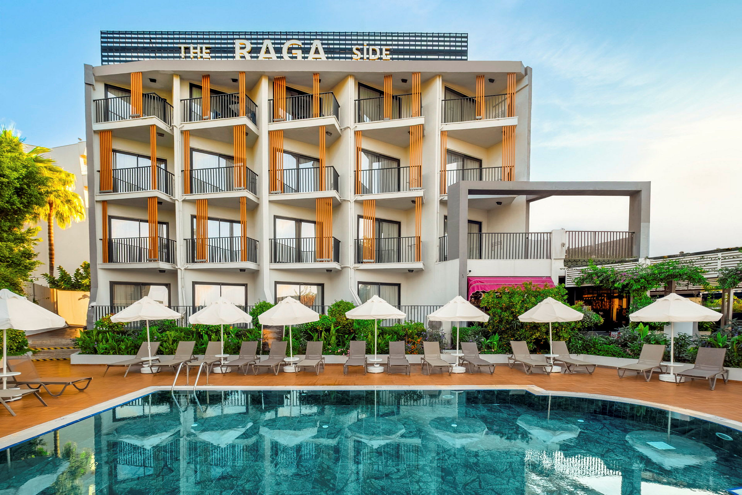 Ett hotell byggt i moderna linjer med en utomhuspool framför, omgiven av solstolar och parasoll. (AI-genererad text)