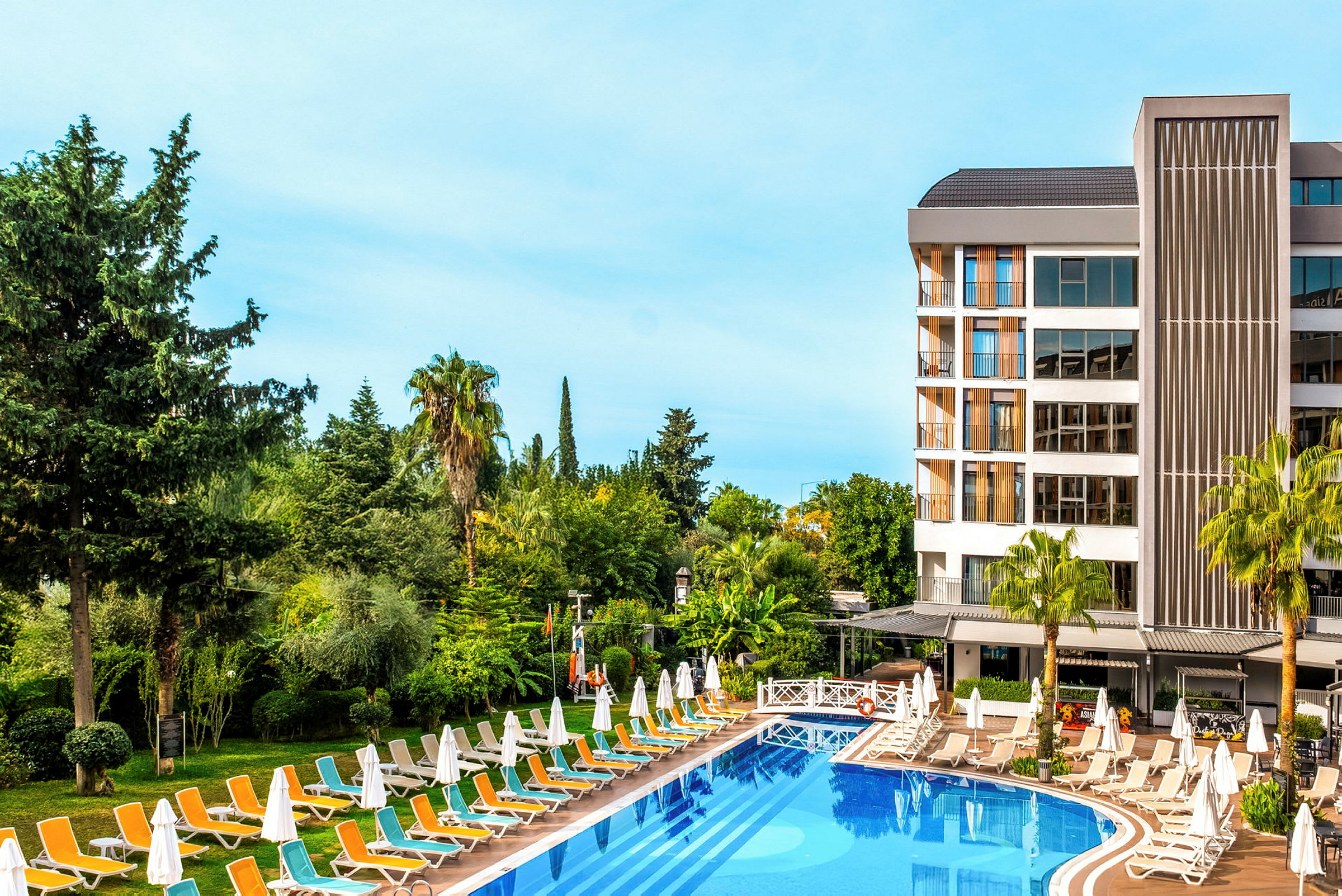 En vy över en pool vid ett hotell omgivet av gröna träd och växtlighet. (AI-genererad text)