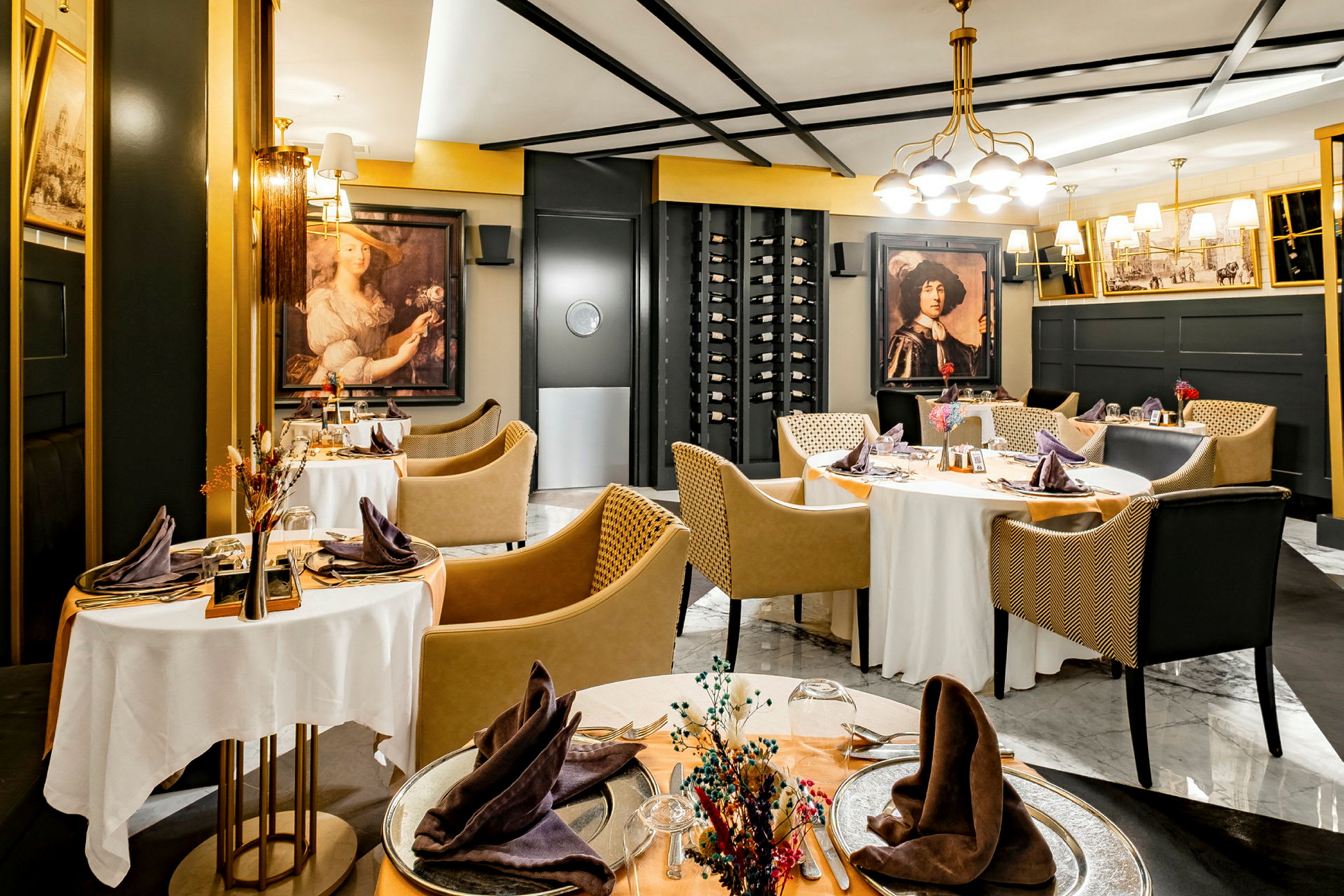 Ett elegant inrett restaurangutrymme med runda bord klädda med vita dukar, omgivna av moderna stolar och dekorationer som inkluderar konstverk på väggarna och en vinhylla. (AI-genererad text)