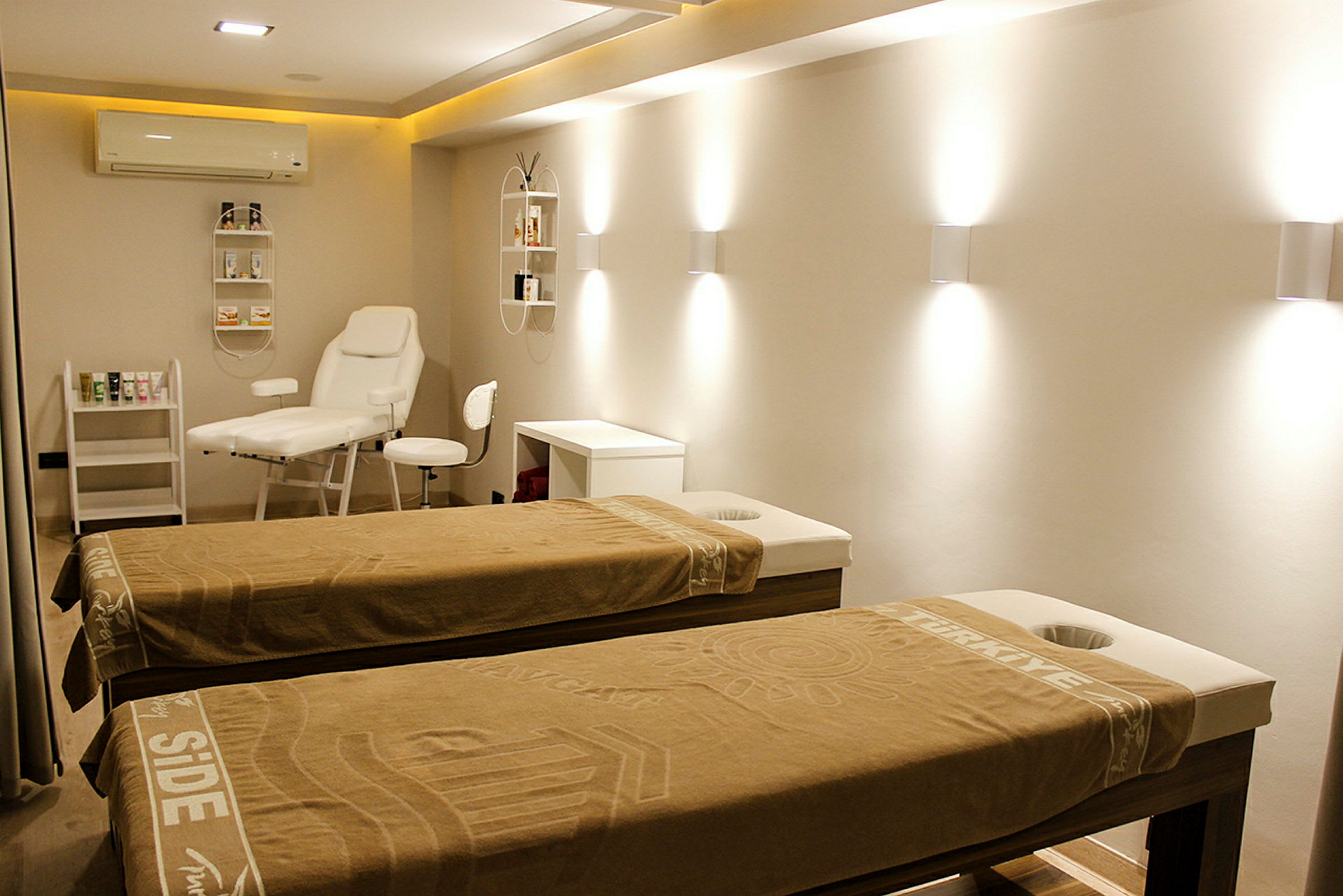 Rummet visar en upplyst och modern behandlingslokal med två massagebänkar täckta med bruna handdukar. På väggen finns hyllor med produkter och en vit behandlingsstol och bord. (AI-genererad text)