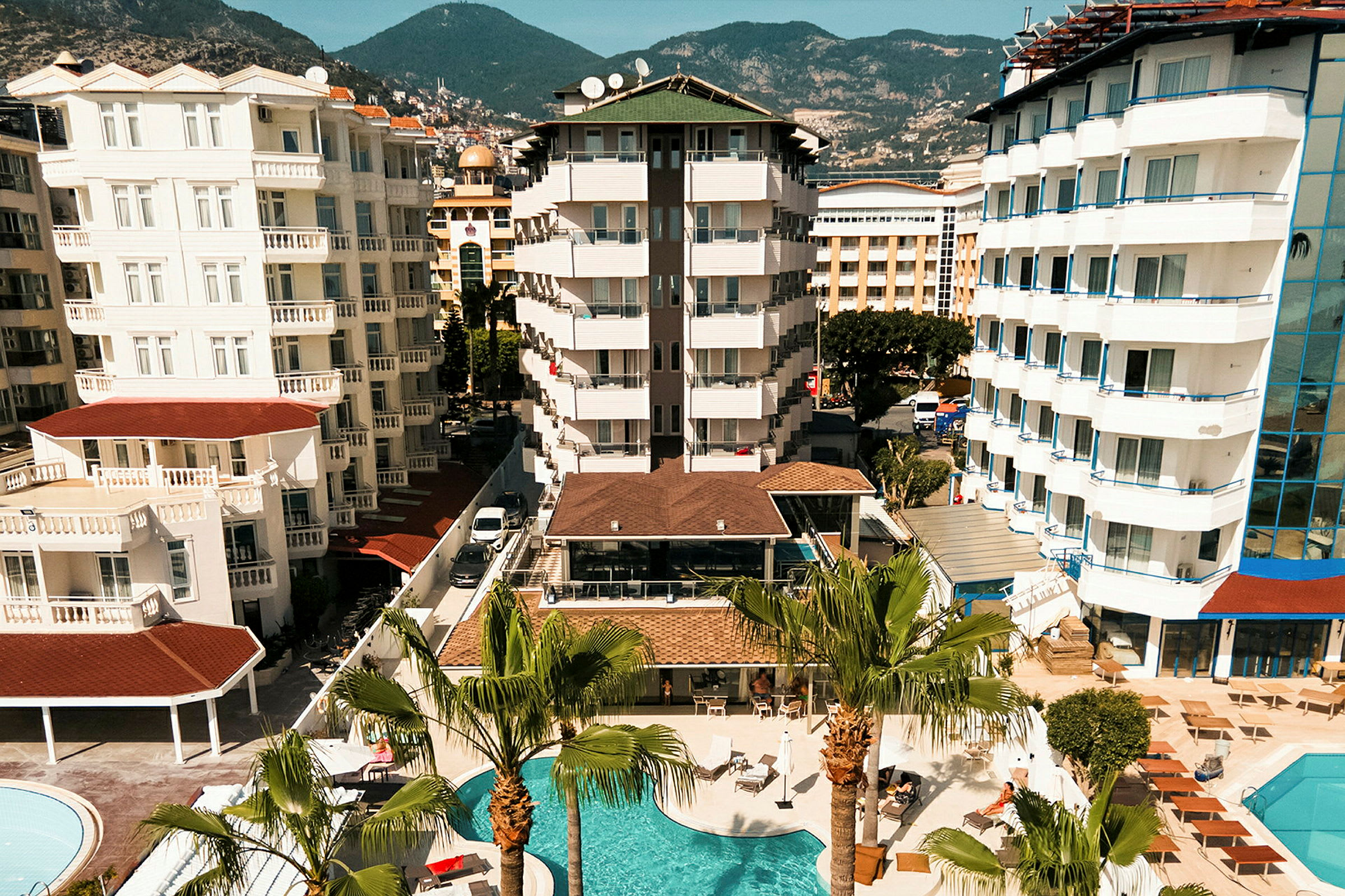 Vy över ett hotellområde med flera byggnader, balkonger och en swimmingpool omgiven av palmer. (AI-genererad text)
