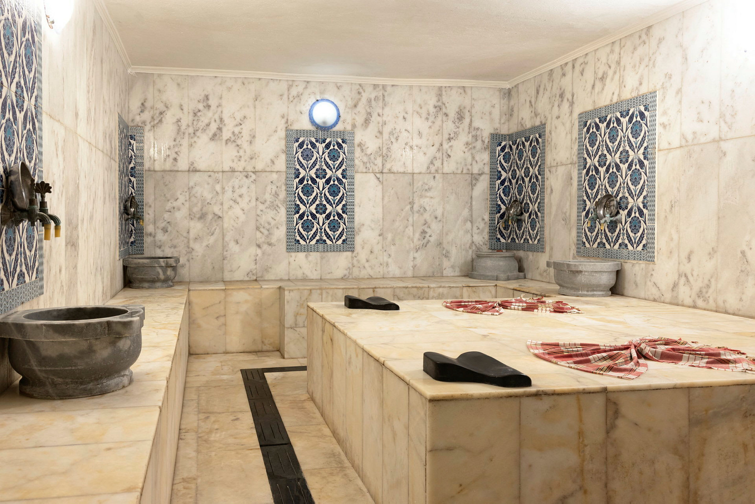 En traditionell hamam med marmorytor och dekorativa kakelplattor på väggarna. Stengropar och kranar syns runtom. (AI-genererad text)
