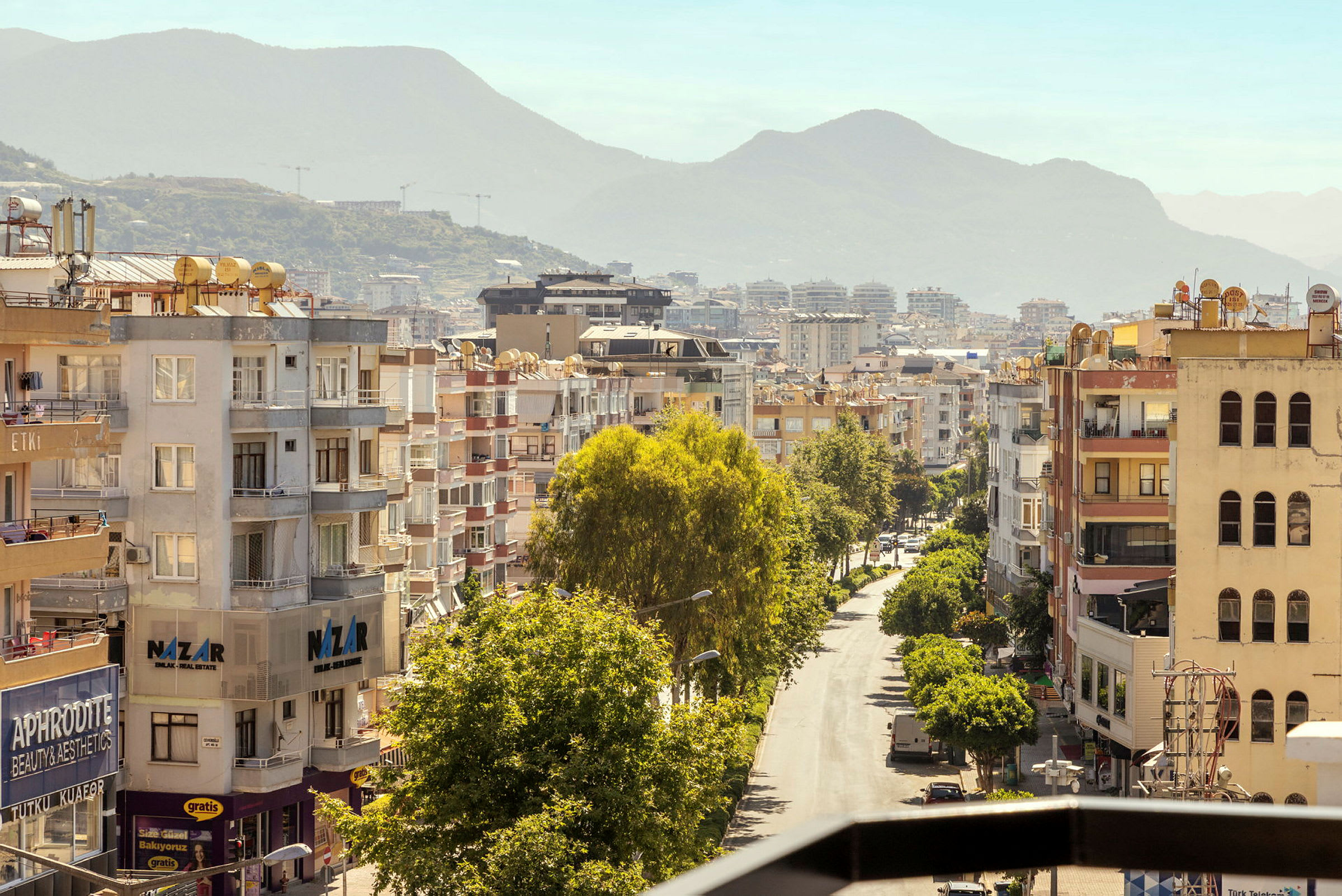 Vy över en stadsgata kantad av flerfamiljshus och grönskande träd, med berg i bakgrunden. (AI-genererad text)