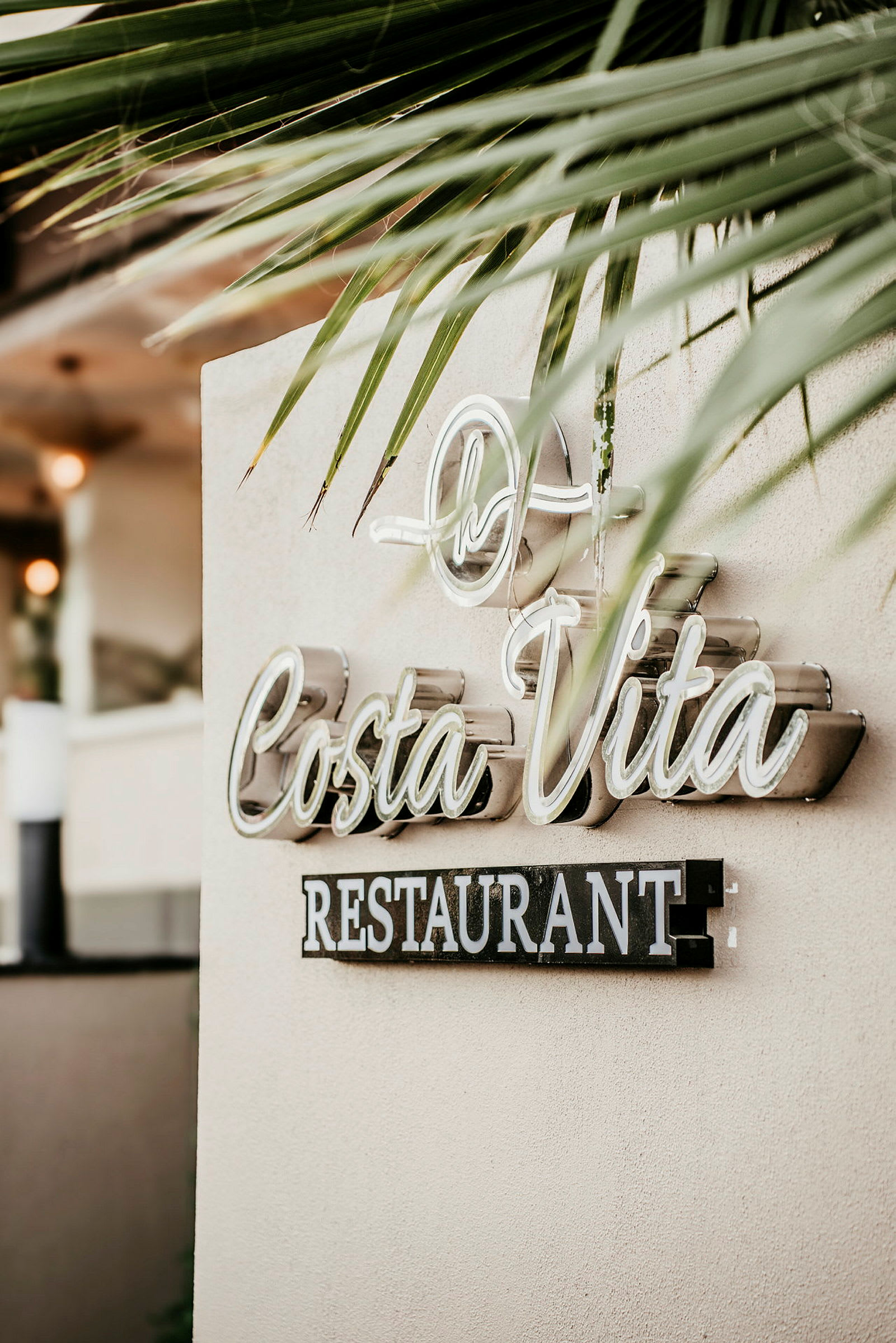 Ett tecken med texten 'Costa Vita Restaurant' på en vägg, omgivande palmbasadekoration. (AI-genererad text)