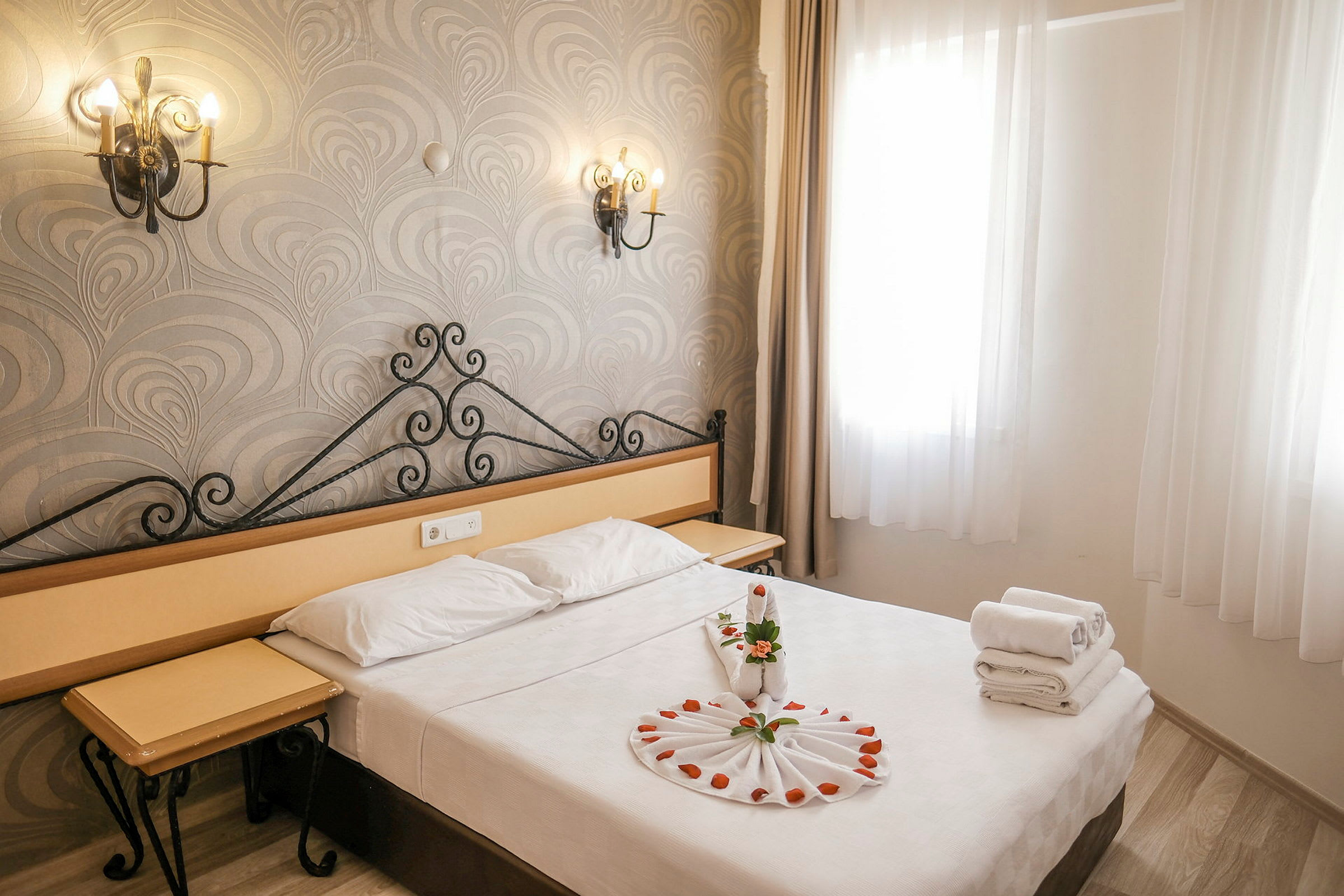 Ett hotellrum med en dubbelsäng, dekorerat med handdukar och blomblad på sängen. Vita gardiner täcker fönstret till höger. (AI-genererad text)