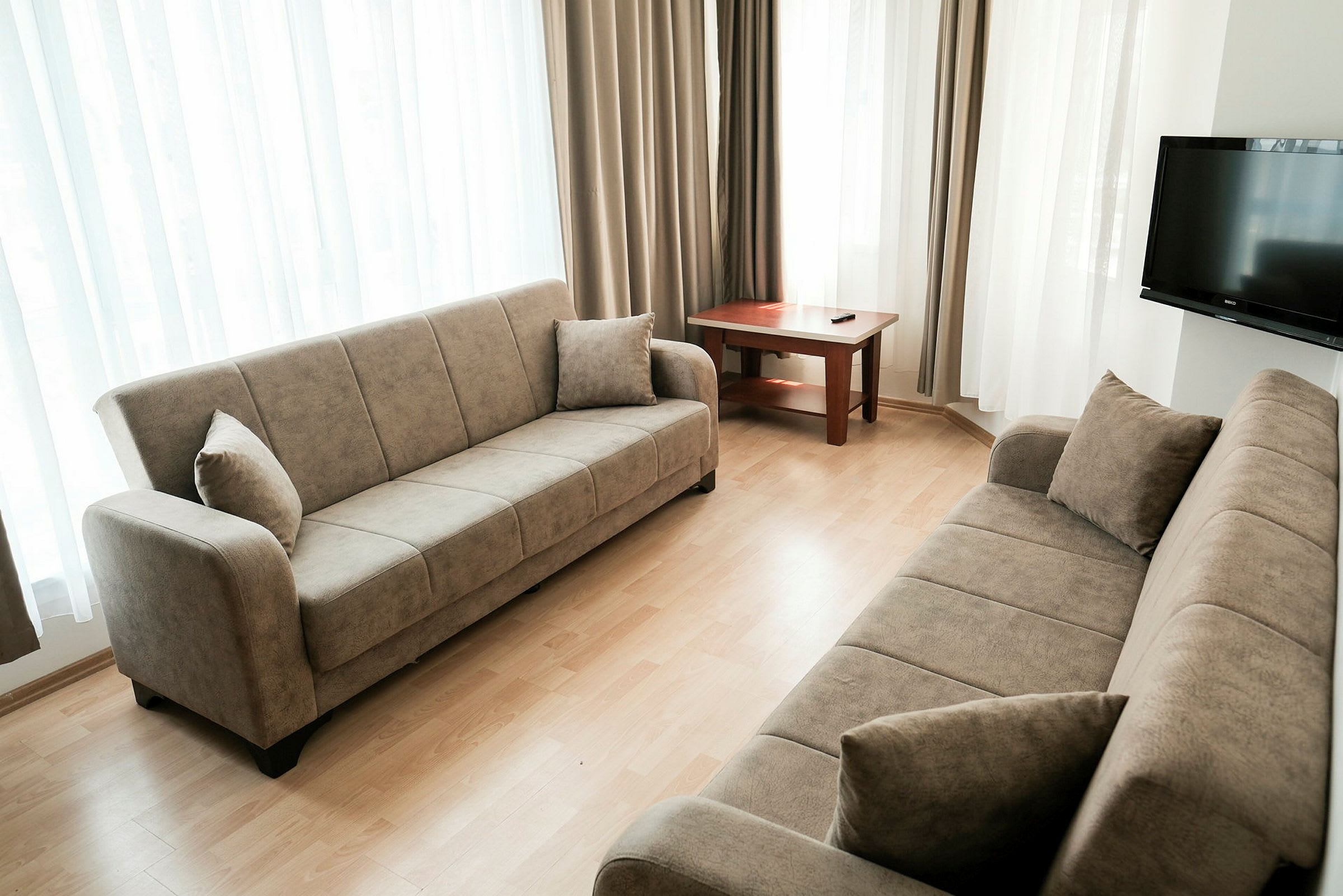 Ett vardagsrum med två beige soffor, ett träbord och en TV på väggen. Rummet har ljusa gardiner vid fönstren. (AI-genererad text)