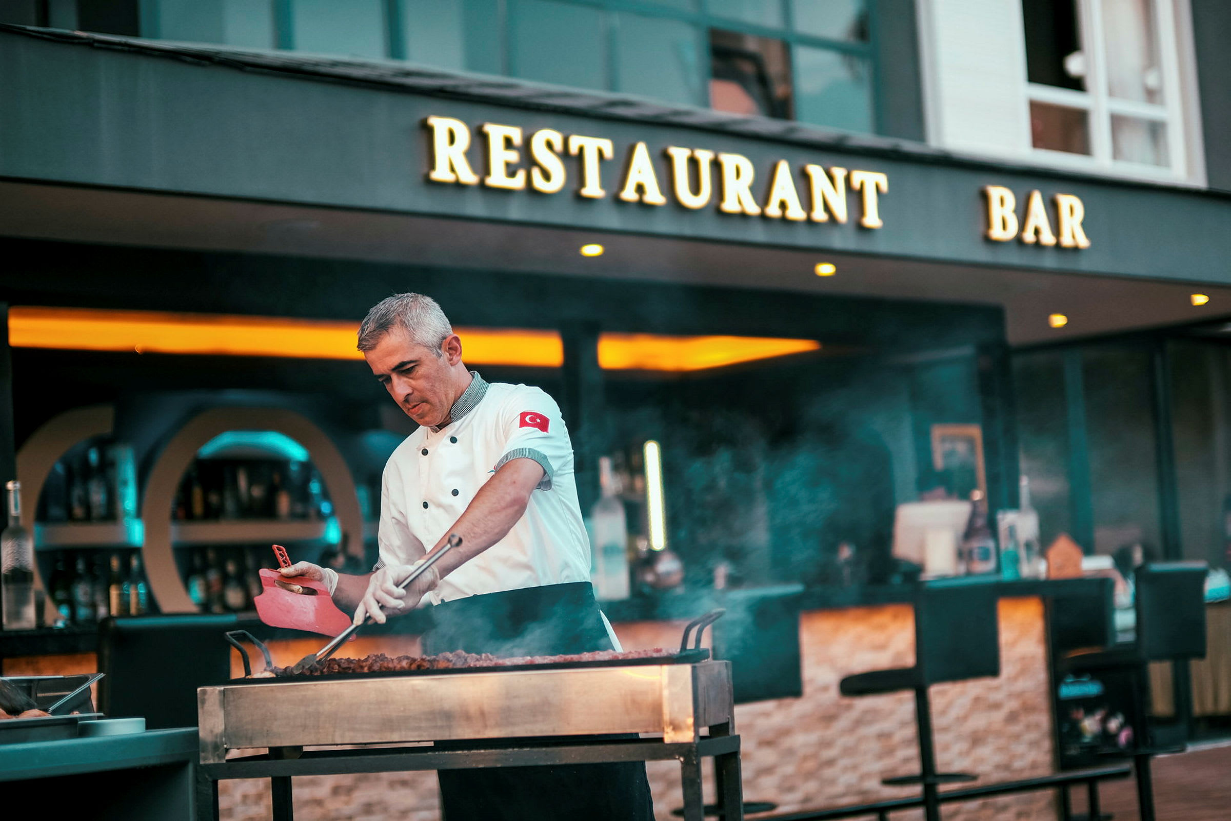 En kock tillagar mat på en öppen grill framför en restaurang och bar. (AI-genererad text)