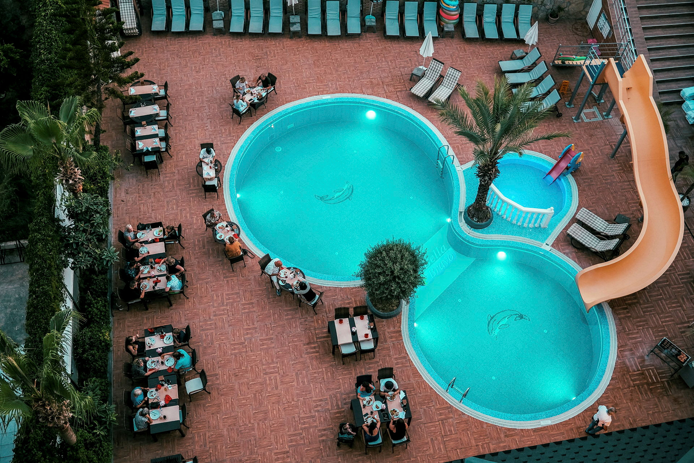 En utomhuspool på ett hotellområde med solstolar och personer som sitter vid bord och äter. (AI-genererad text)