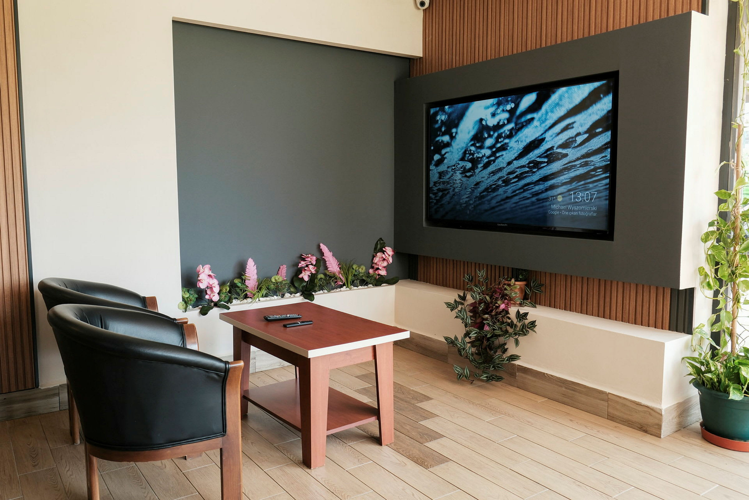 Ett rum med två svarta fåtöljer, ett träbord och en stor väggmonterad TV. Det finns dekorativa blommor och växter runtom. (AI-genererad text)