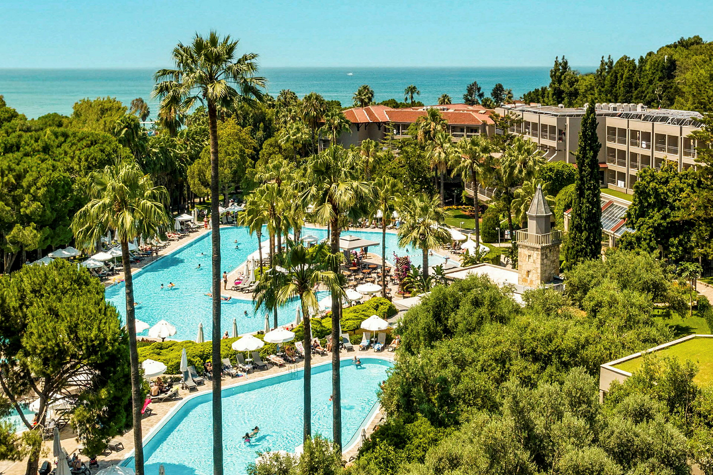 Ett hotellområde med pooler omgivna av palmer och grönska, i bakgrunden syns havet. (AI-genererad text)