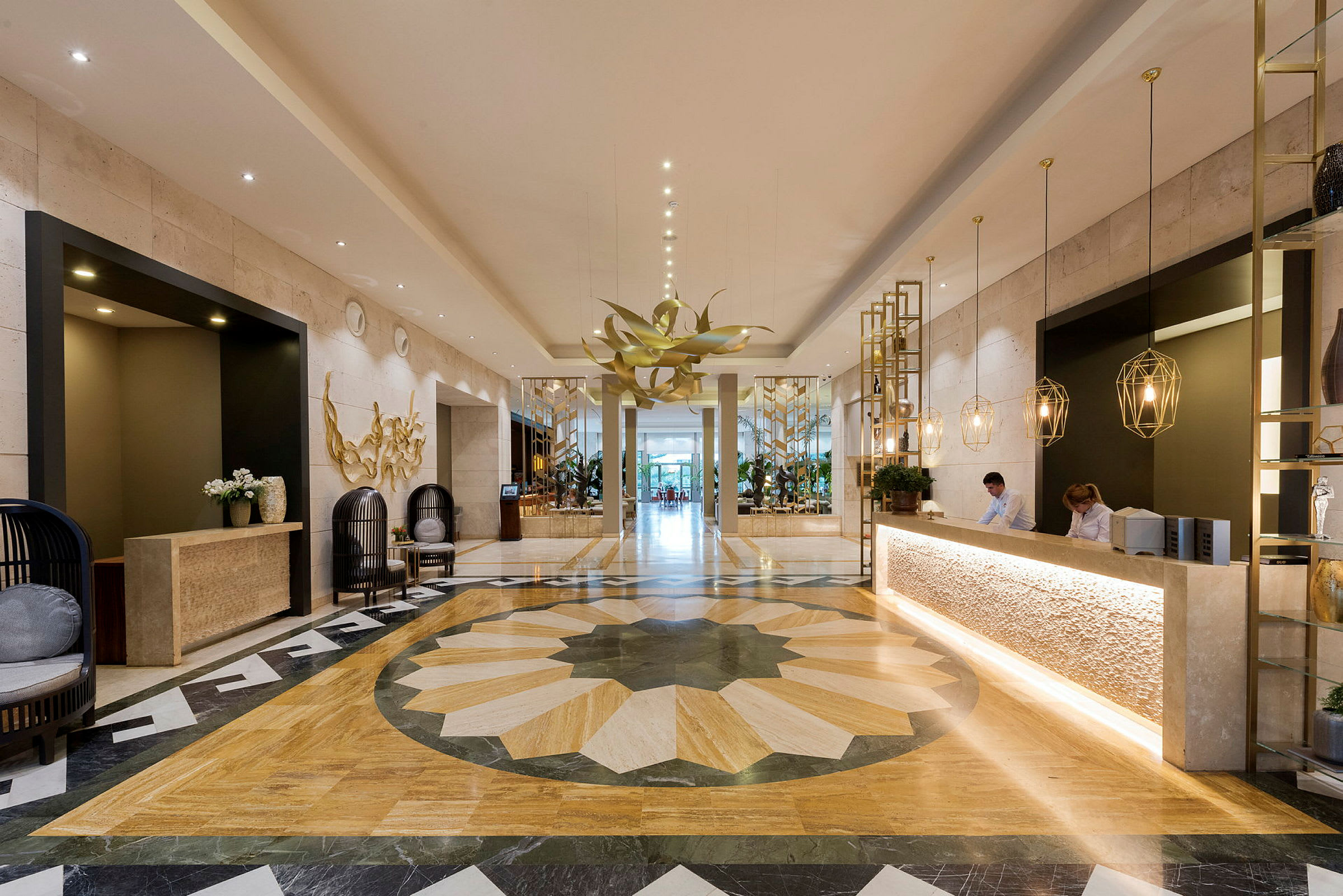 Ett elegant hotellobby med marmorgolv, en reception med anställda, moderna dekorationer och en stor blomliknande ljuskrona. (AI-genererad text)
