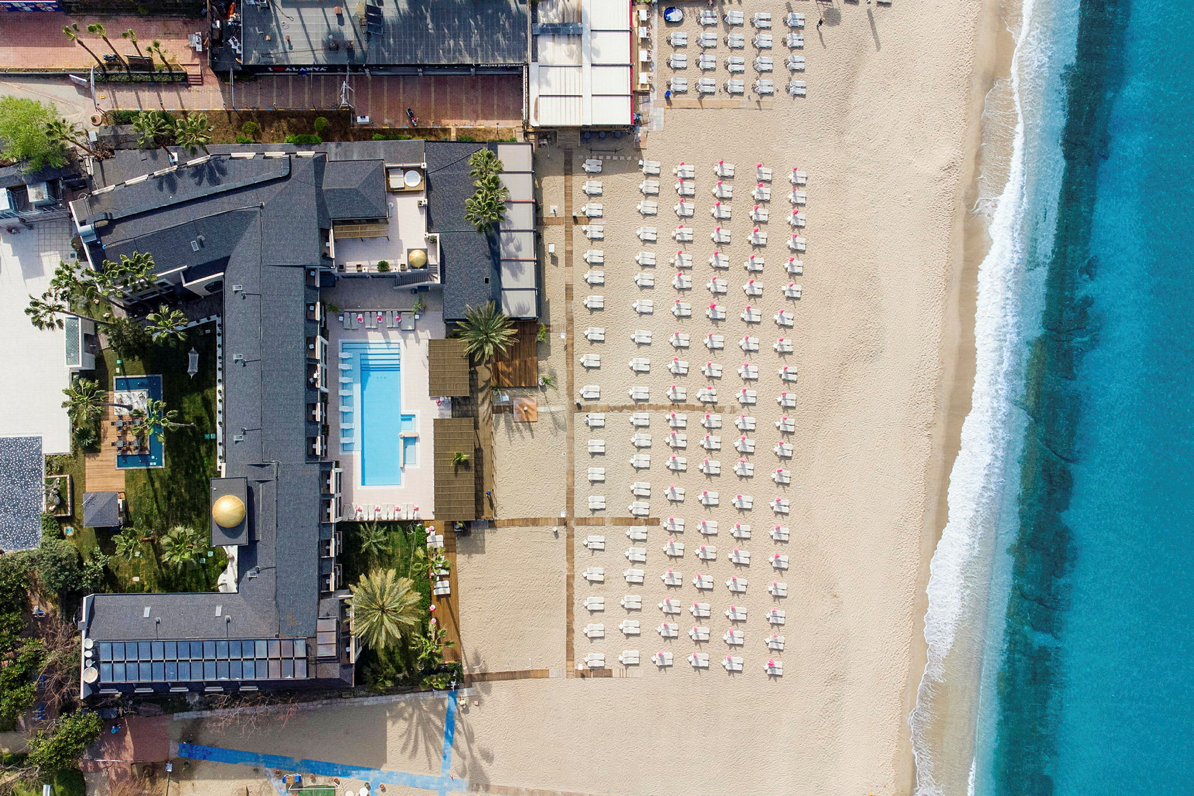 Flygvy över en strand med solstolar, ett hotellområde med pool, och havet. (AI-genererad text)