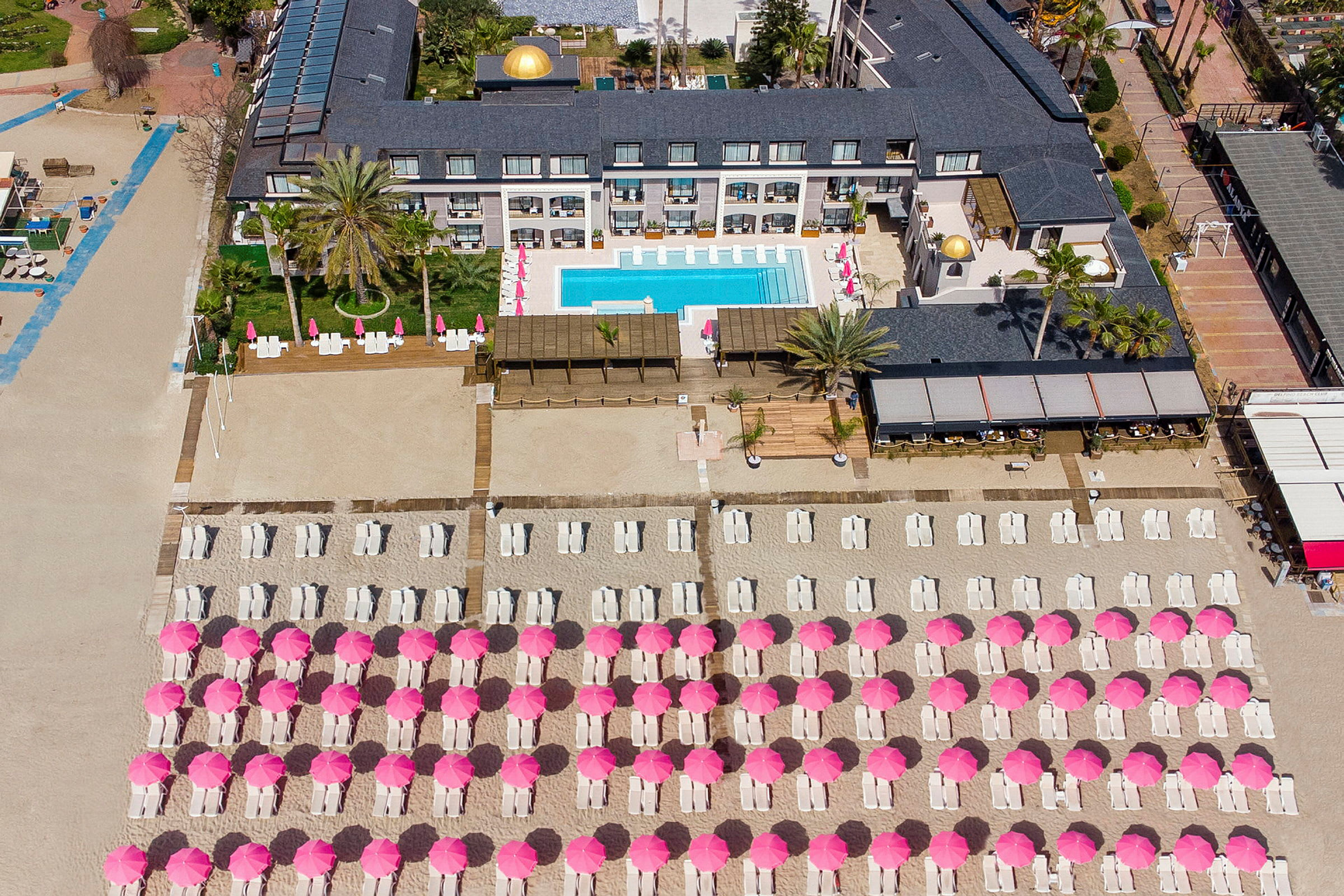 Fågelsikt över hotellområde med pool och solstolar under rosa parasoller på stranden. (AI-genererad text)