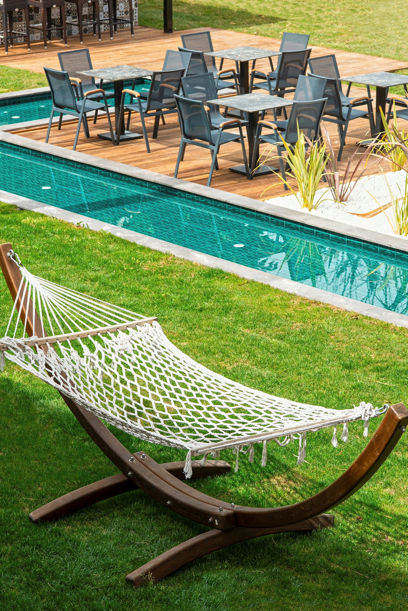 En hängmatta av nät på en gräsmatta intill en pool med sittmöbler på en träplattform. (AI-genererad text)