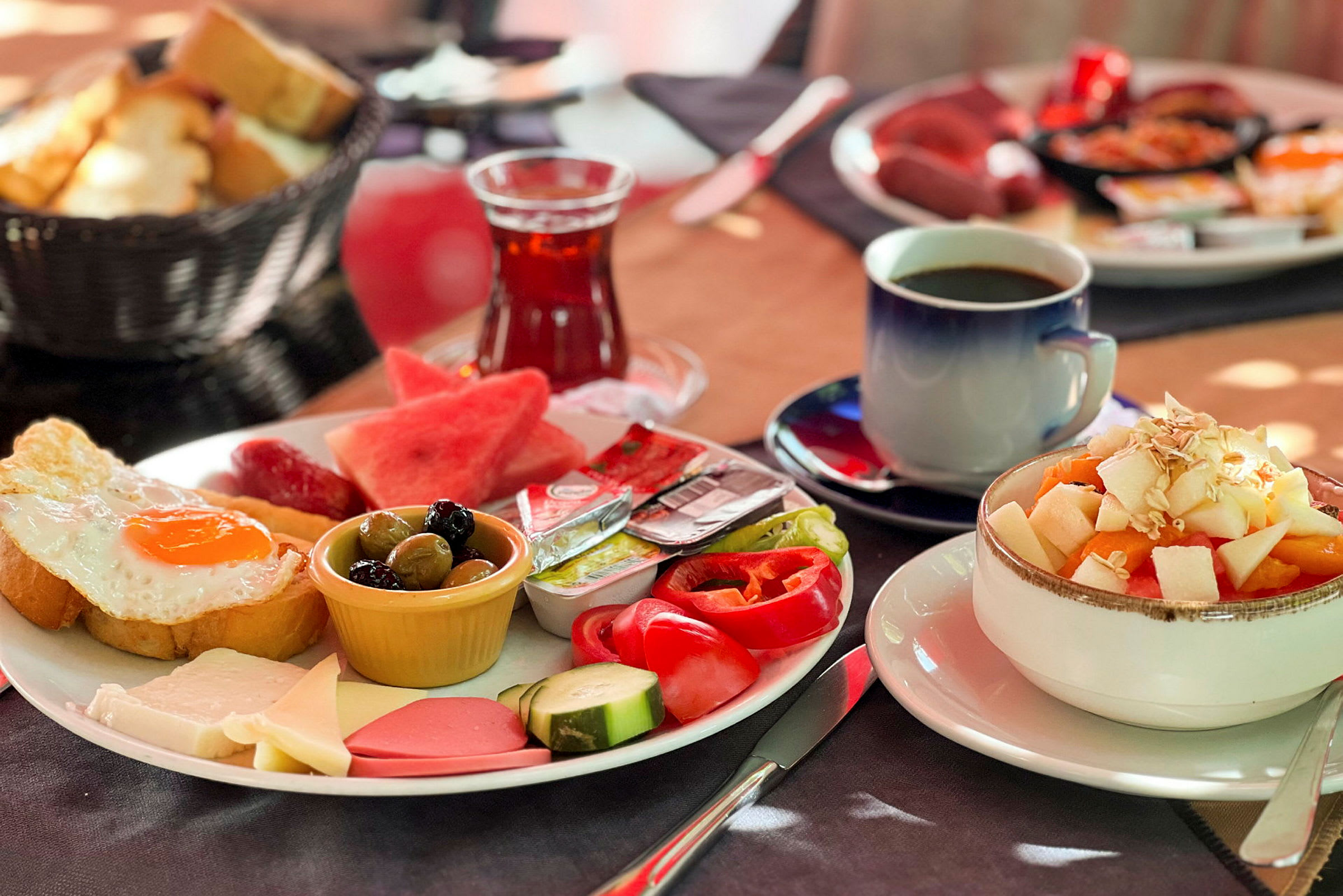 En tallrik med frukostmat inklusive stekt ägg, grönsaker, ost och andra tillbehör, bredvid en skål med fruktsallad och drycker. (AI-genererad text)