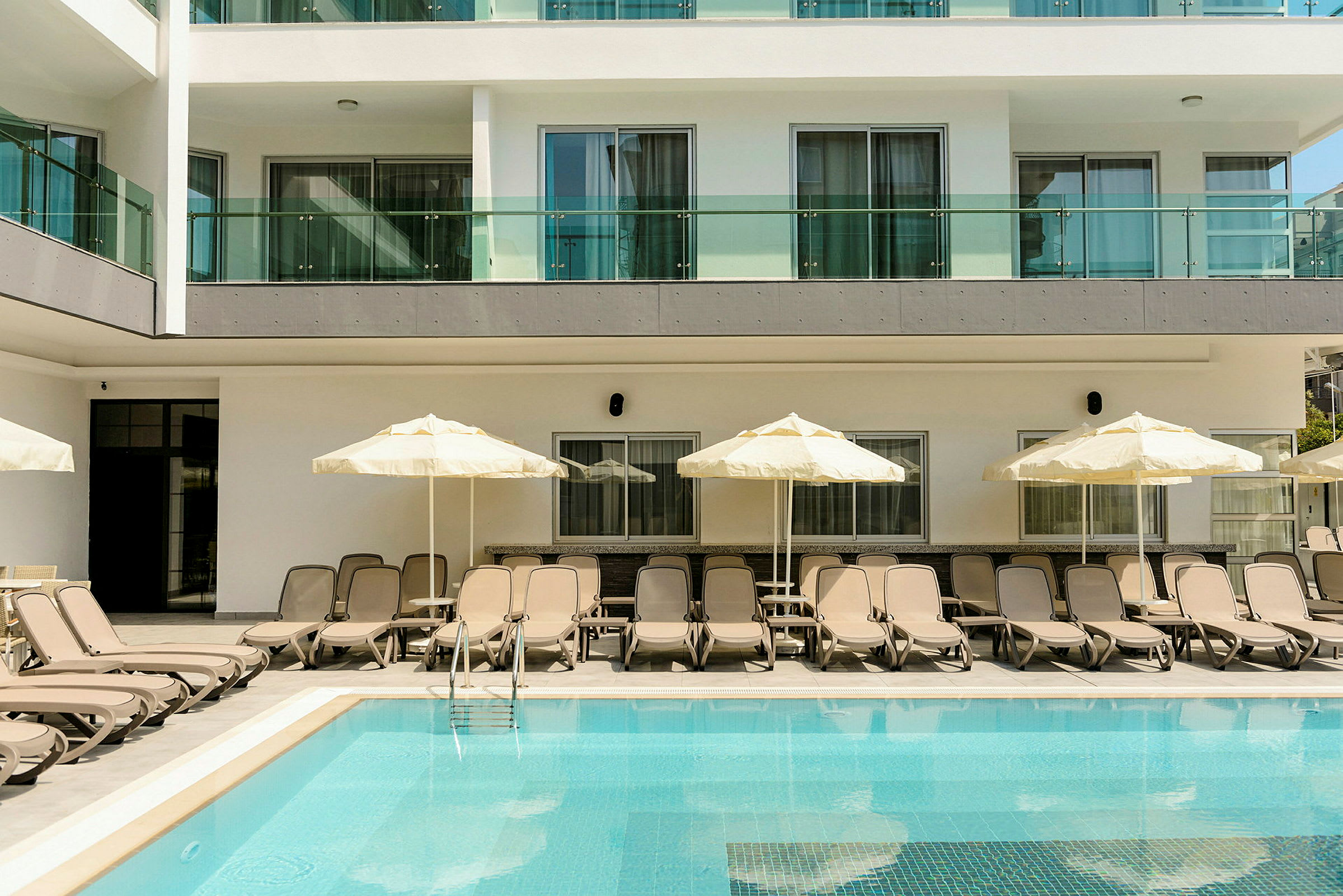 En pool med solstolar och parasoller vid sidan, framför en modern byggnad. (AI-genererad text)