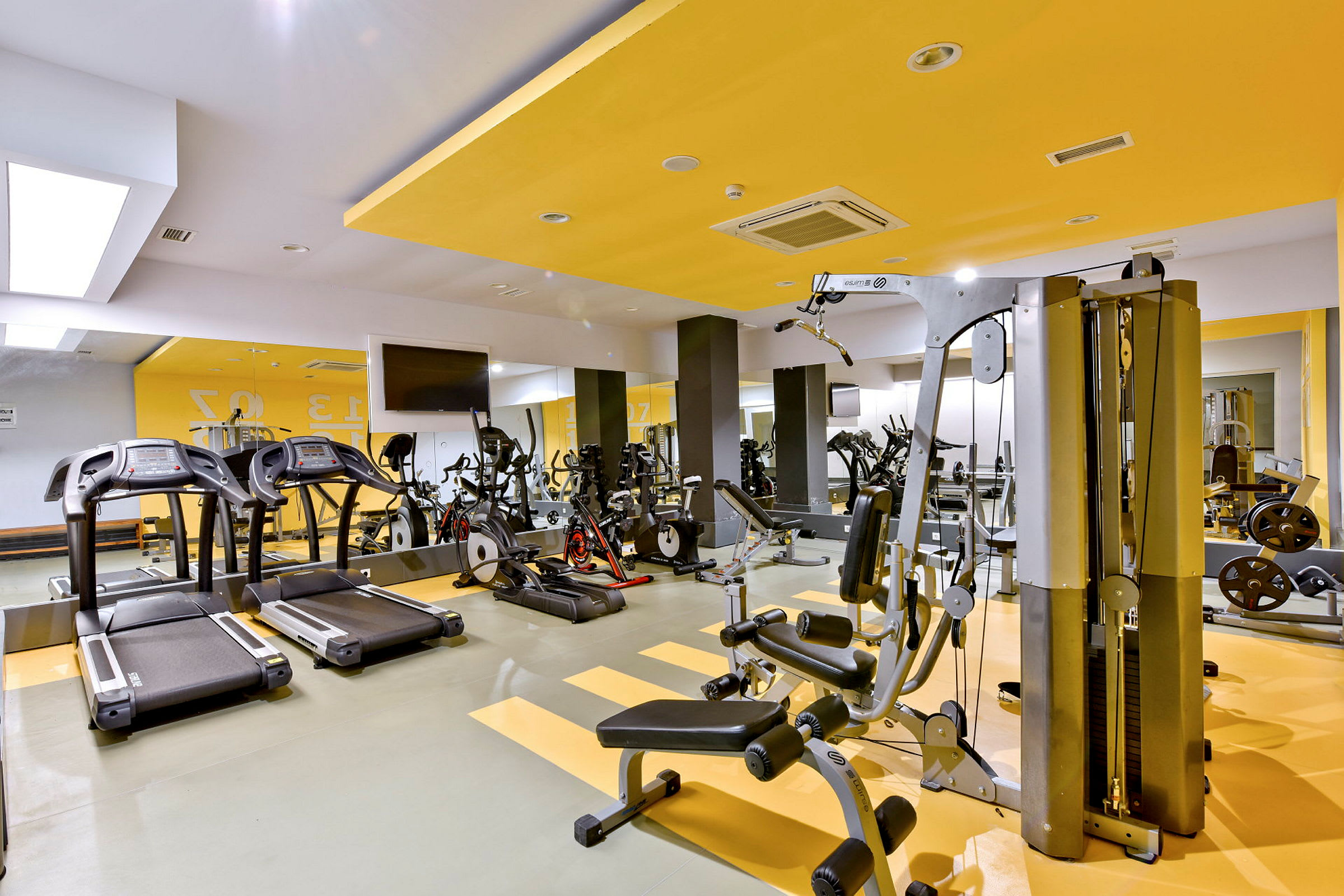 Ett gym med olika träningsmaskiner såsom löpband, cyklar och styrketräningsutrustning. Väggarna är målade i gult och vitt. (AI-genererad text)