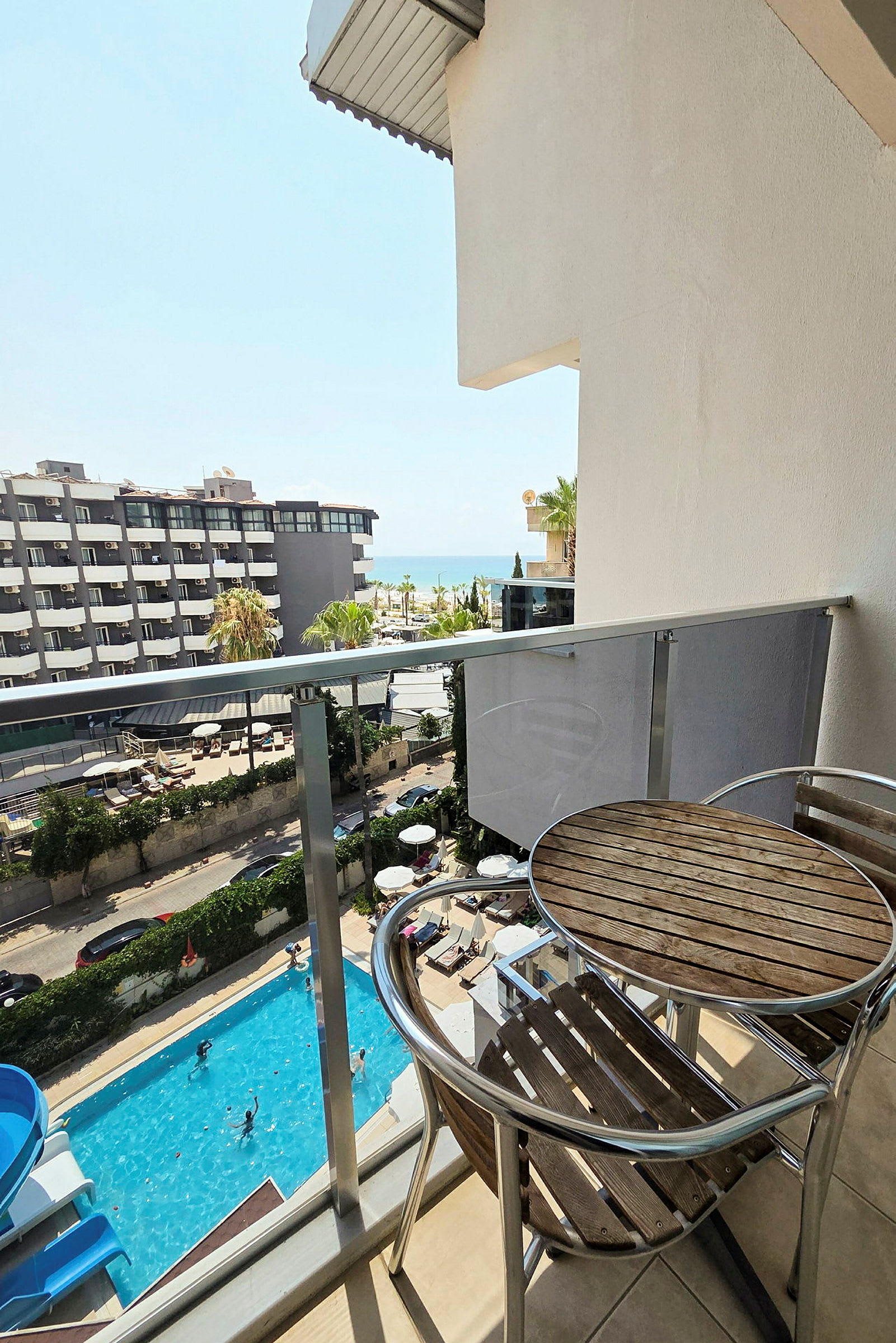 En balkong med ett bord och två stolar. Utanför syns en swimmingpool, parasoller och en byggnad samt havet i bakgrunden. (AI-genererad text)