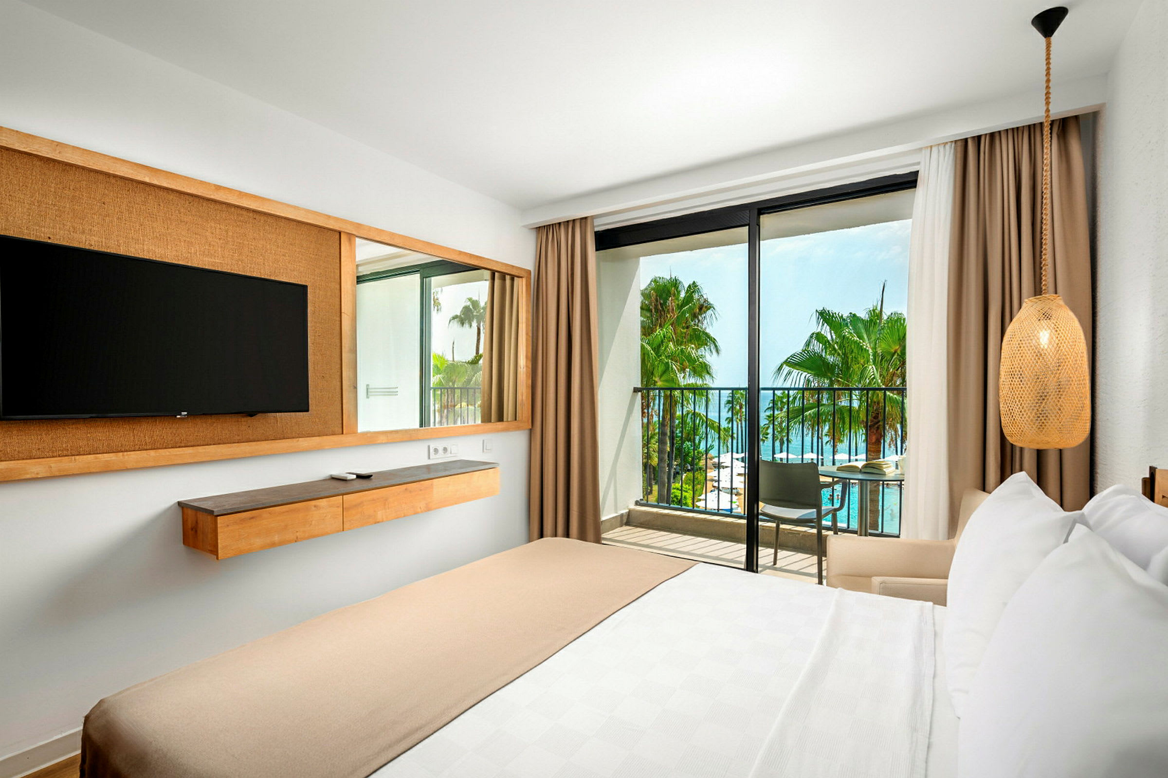 Hotellrum med säng, vägg-TV, balkong och utsikt över pool och palmer. (AI-genererad text)
