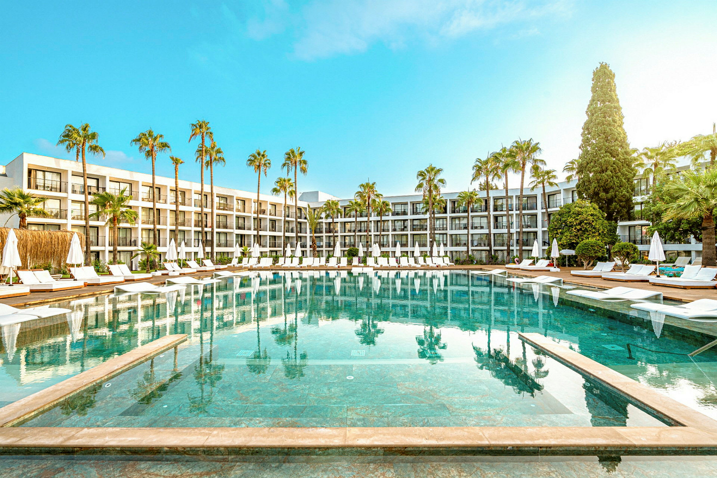 Hotellpool med solstolar och parasoller omgiven av palmträd och moderna byggnader. (AI-genererad text)