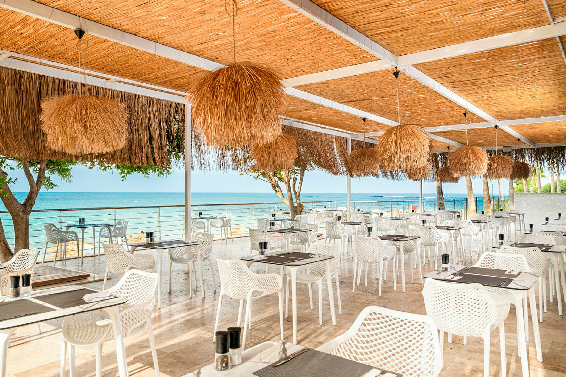 En restaurang med uteservering och havsutsikt, möblerad med vita stolar och bord samt tak dekorerat med strålampor. (AI-genererad text)
