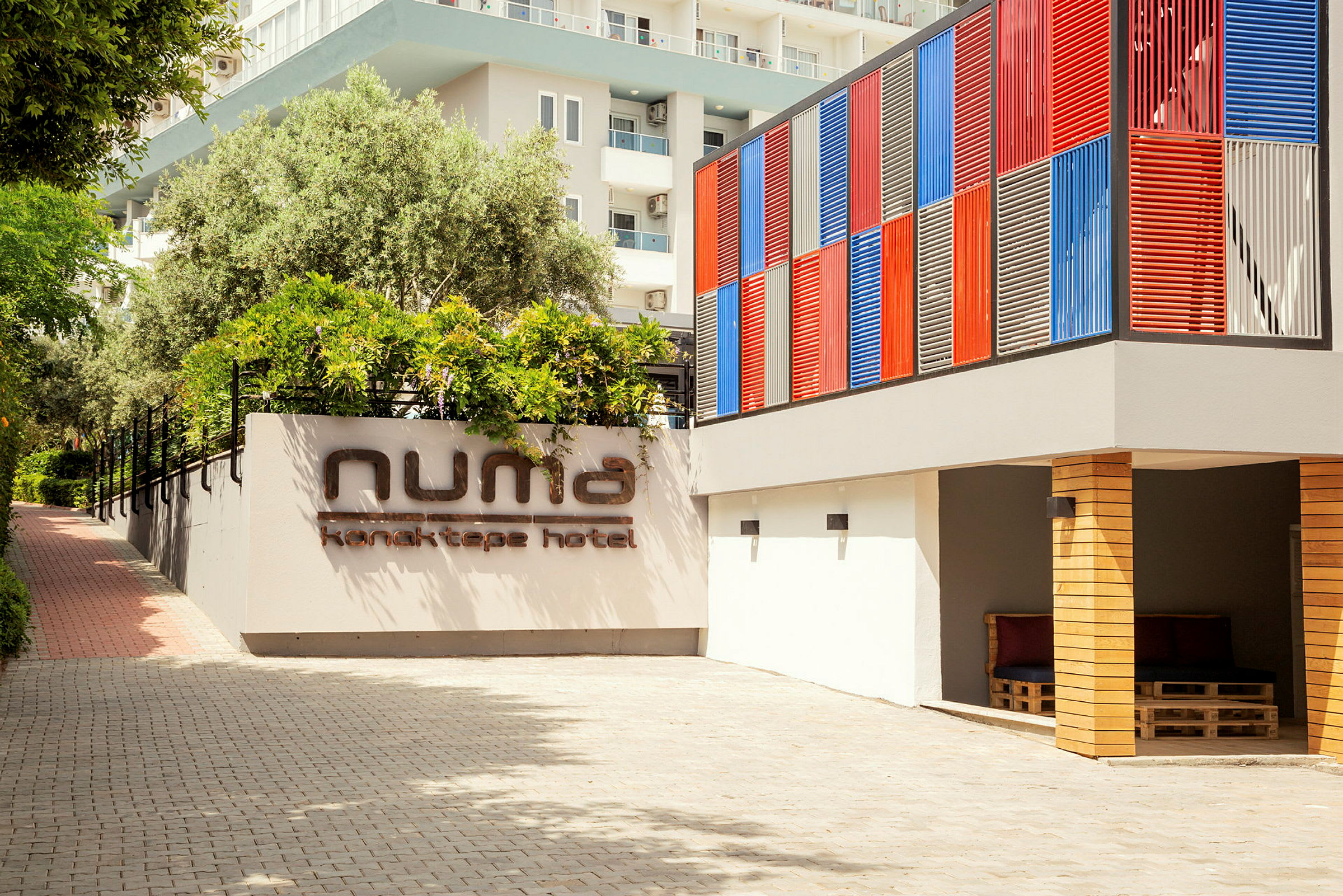 Hotellets fasad med en skylt som säger 'Numa Konaktepe Hotel', omgiven av växter och en färgglad vägg. (AI-genererad text)