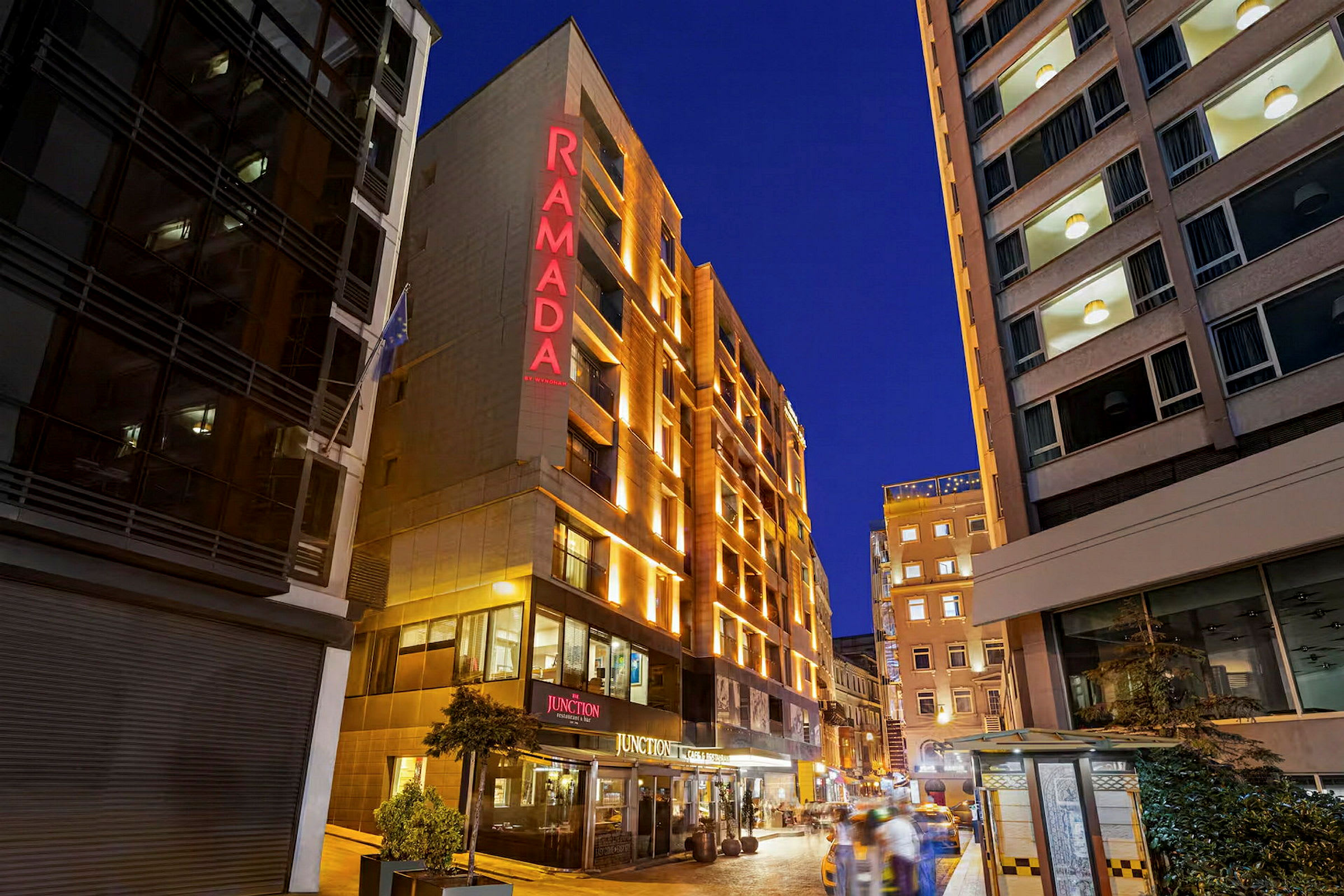 På bilden syns fasaden av ett hotell med texten "RAMADA" belyst i rött, omgiven av andra byggnader och en kvällsupplyst stadsmiljö. (AI-genererad text)