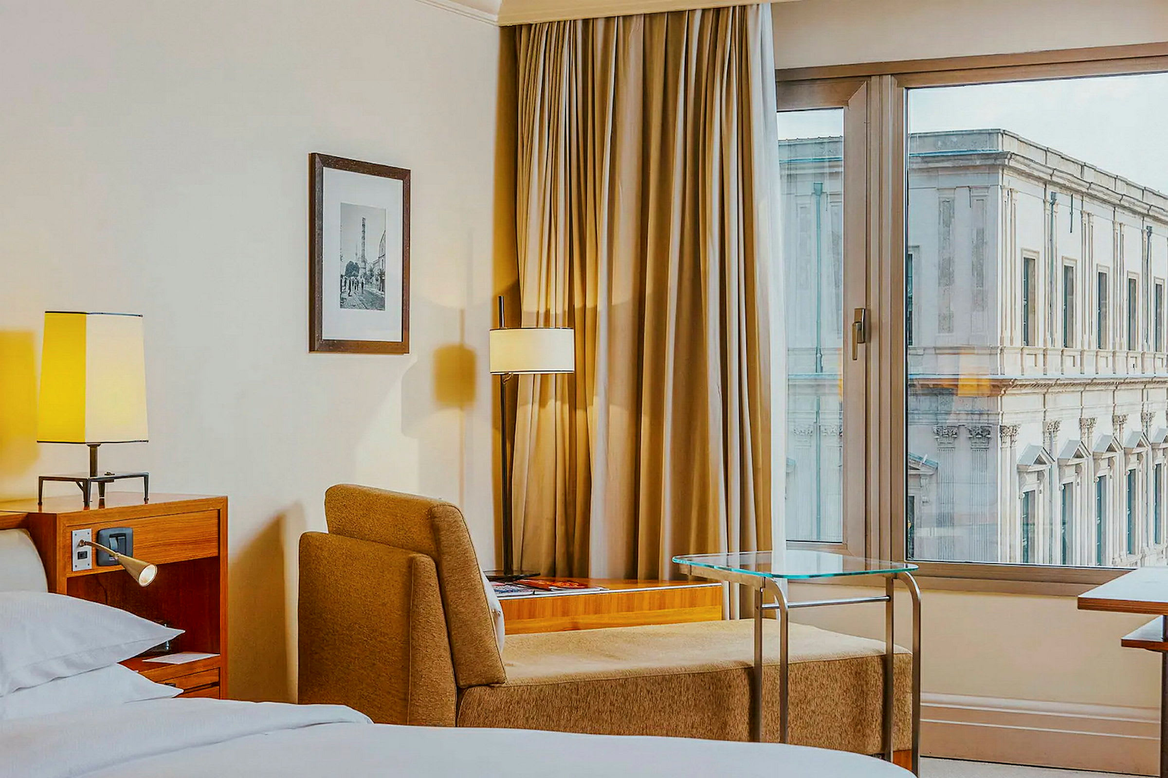 Hotellrum med säng, fåtölj, bord och fönster med utsikt mot en byggnad. (AI-genererad text)