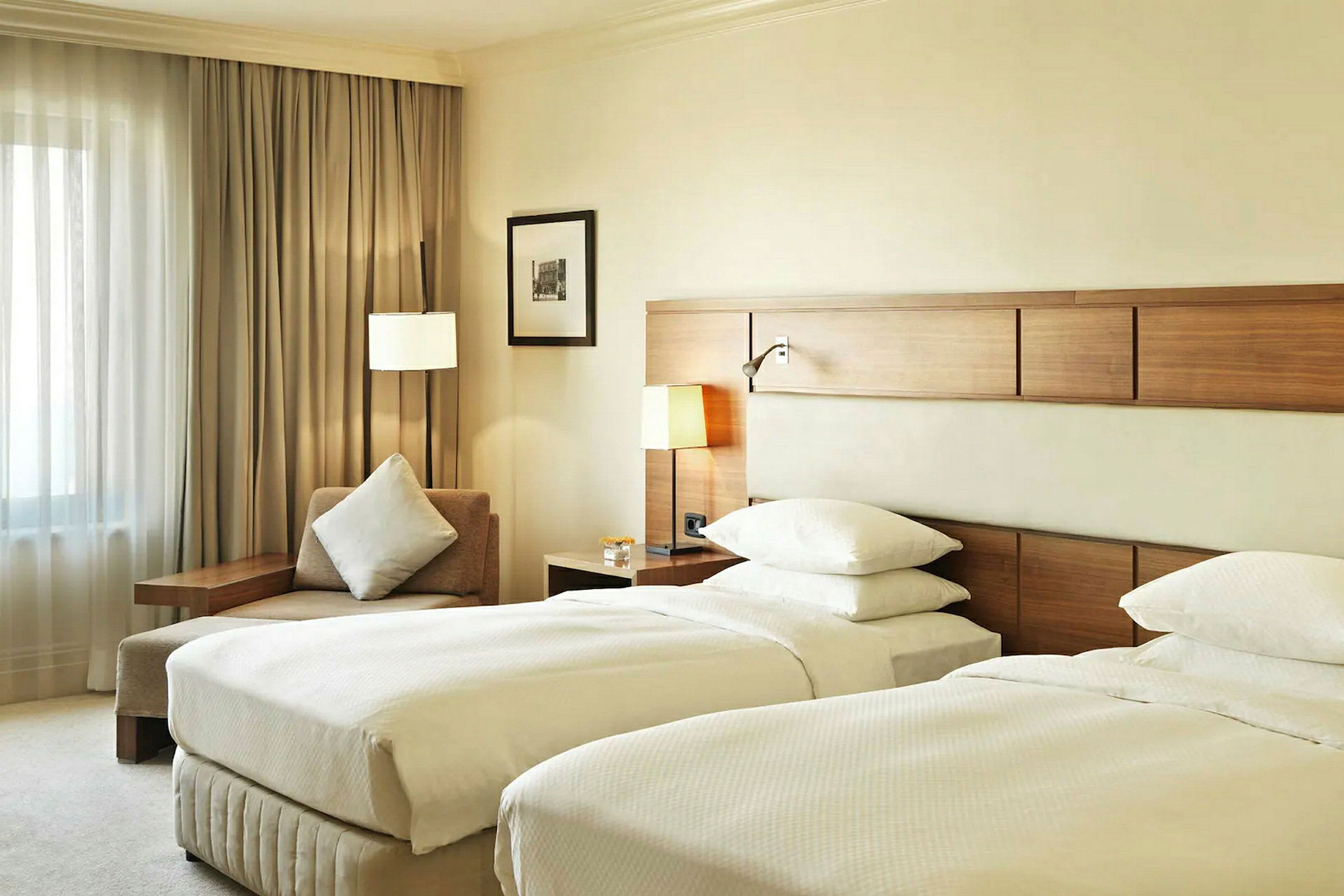 Ett hotellrum med två enkelsängar, en sittplats med kudde och ett fönster med draperier. (AI-genererad text)